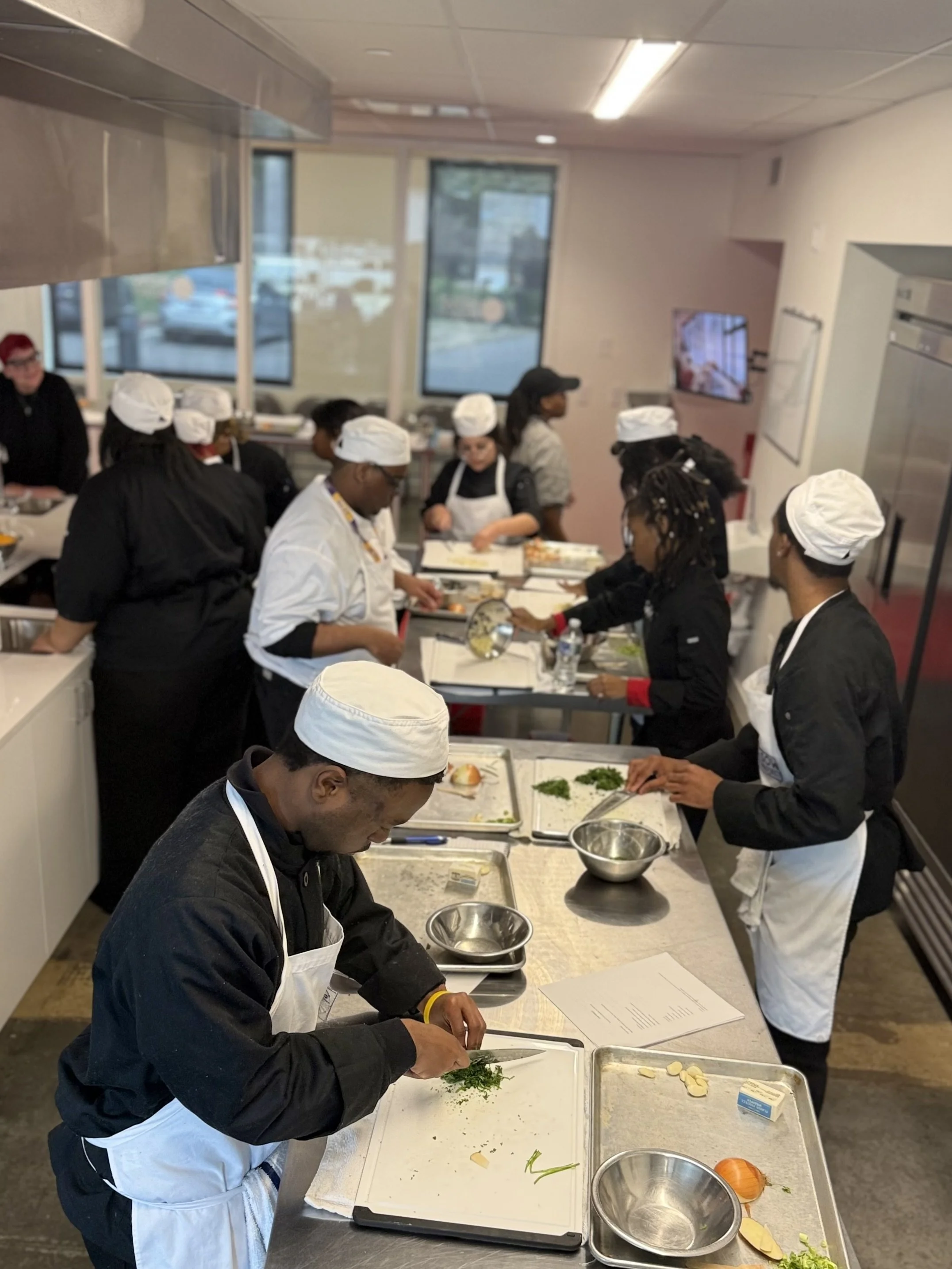 People in a commercial kitchen participating in a cooking class, chopping vegetables, and preparing food.