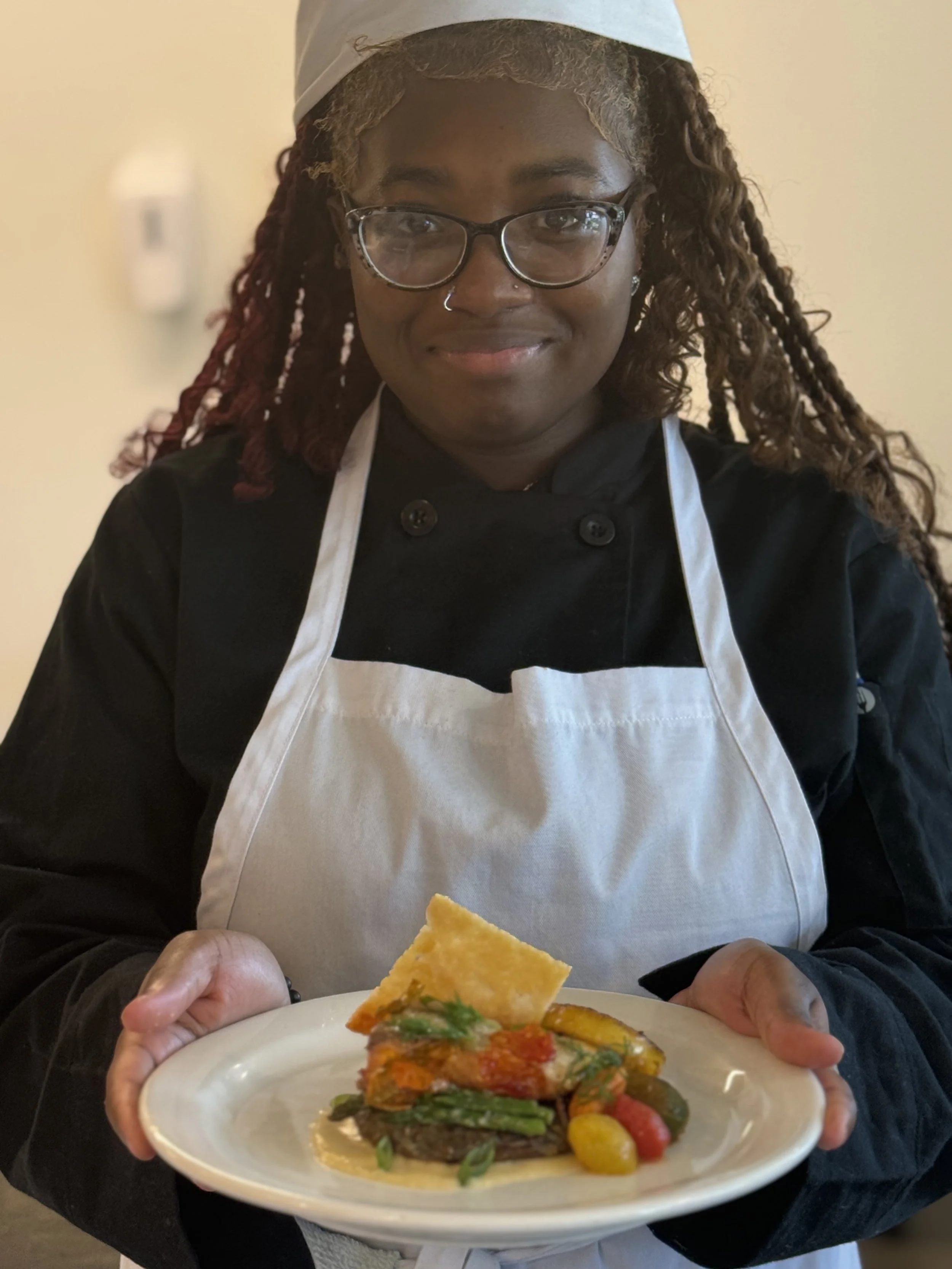 A female chef wearing glasses, a white apron, and a chef's hat, holding a plate with a gourmet dish.