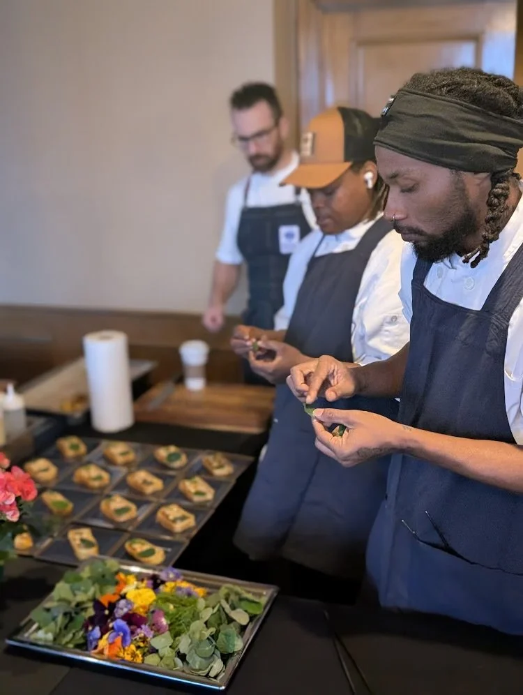 Three chefs, two men and one woman, preparing garnished appetizers on a kitchen counter, with a tray of cocktails and other garnish ingredients in front of them.
