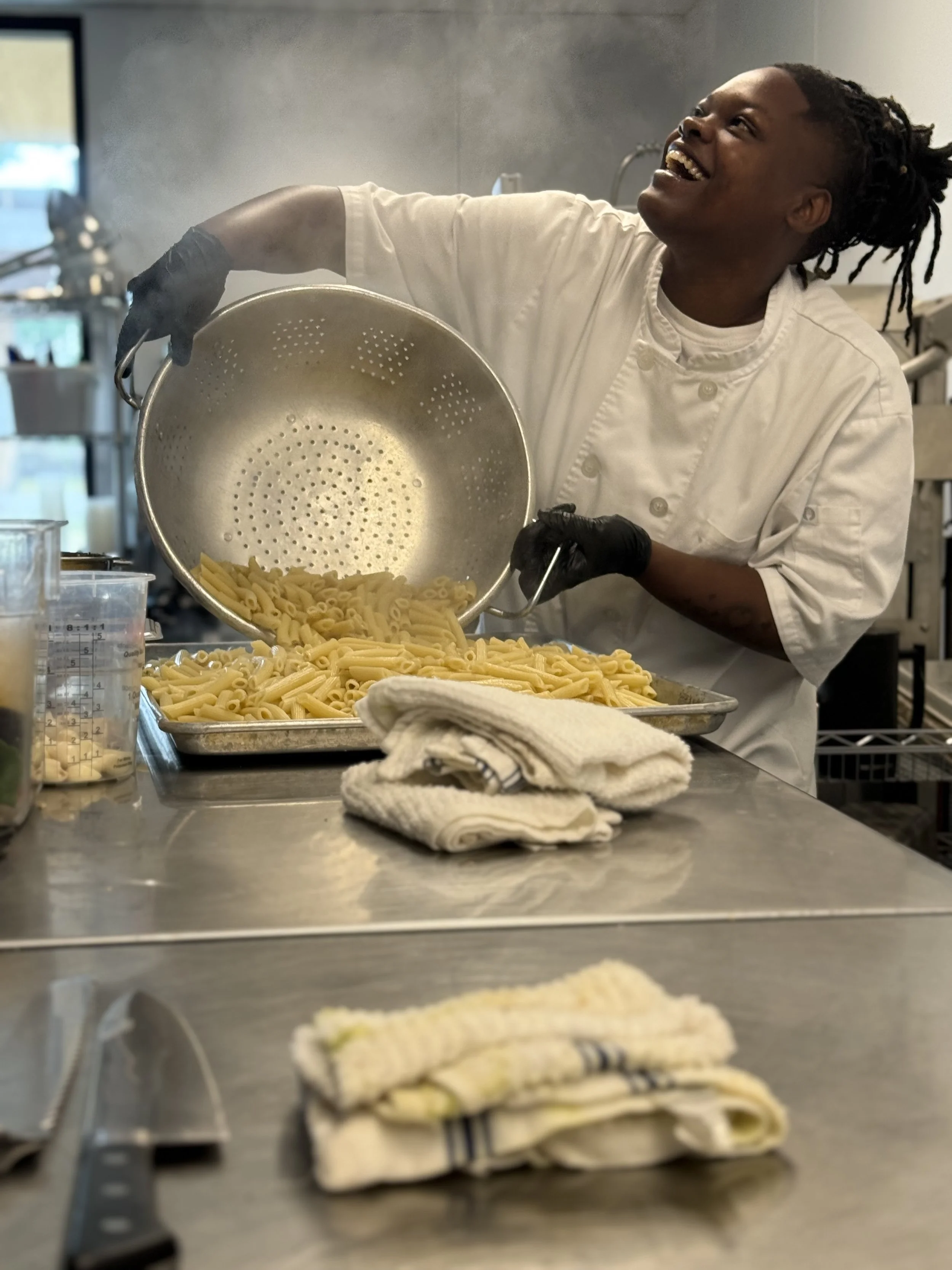A chef in a white uniform and black gloves is smiling and pouring cooked pasta from a colander onto a tray in a professional kitchen.