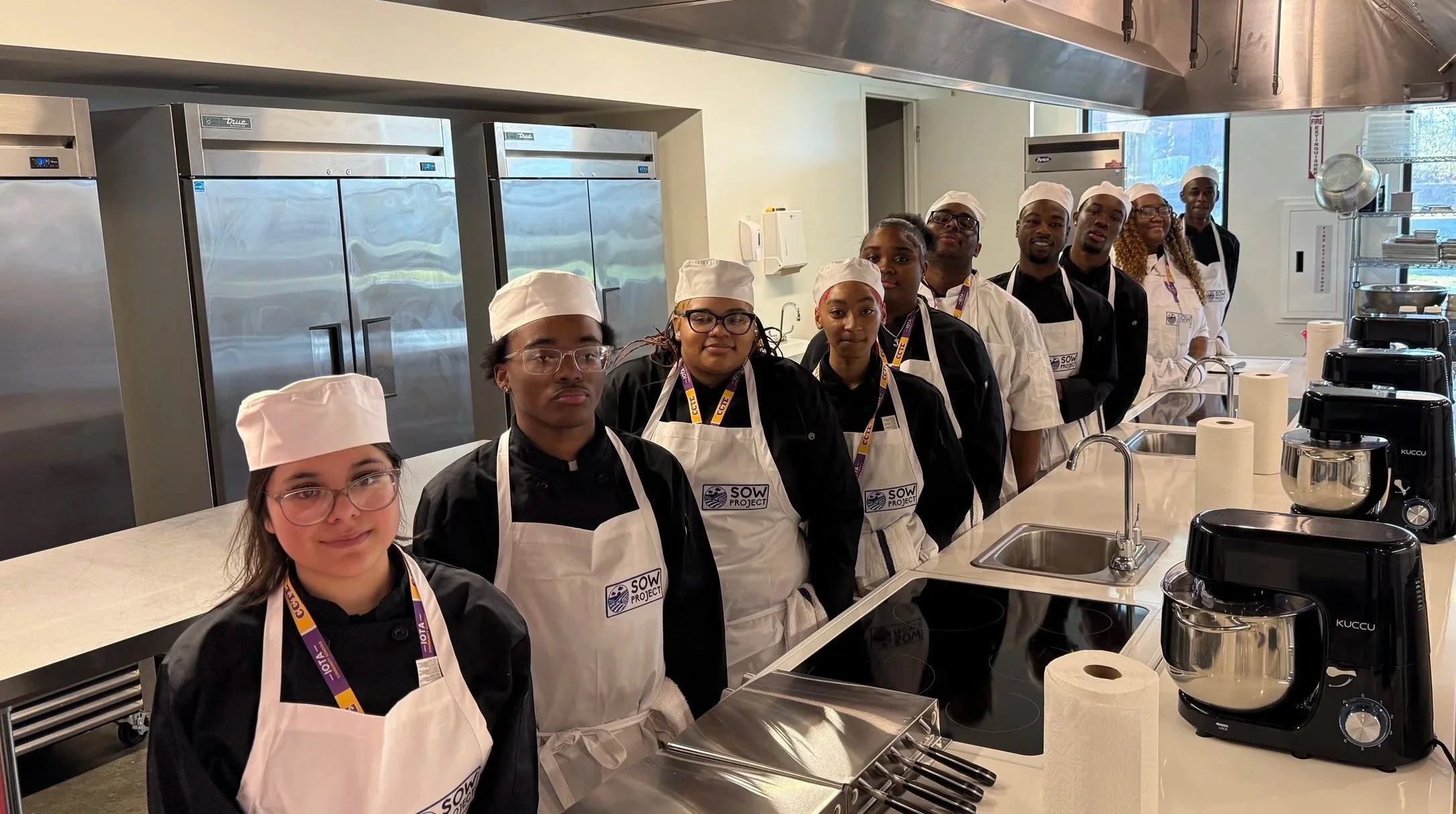 A group of diverse young culinary students standing in a professional kitchen, wearing aprons and chef hats, with various kitchen appliances and stainless steel refrigerators in the background.