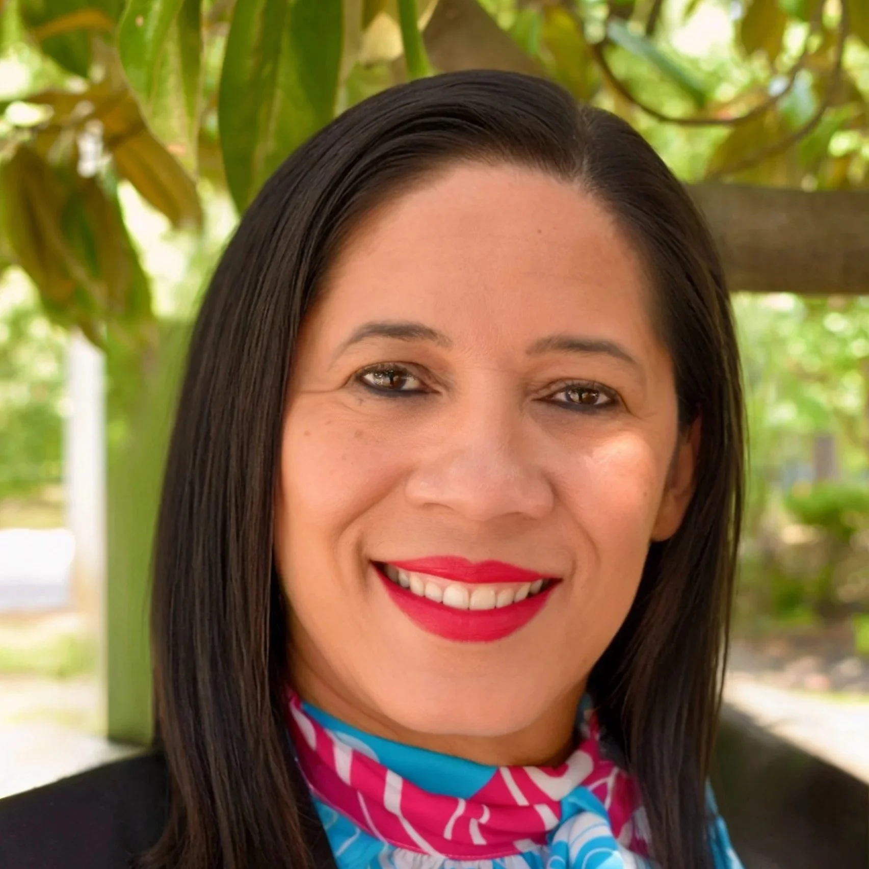 A woman with black hair, wearing a pearl necklace, red lipstick, and a black and white patterned top, smiling against a dark background.