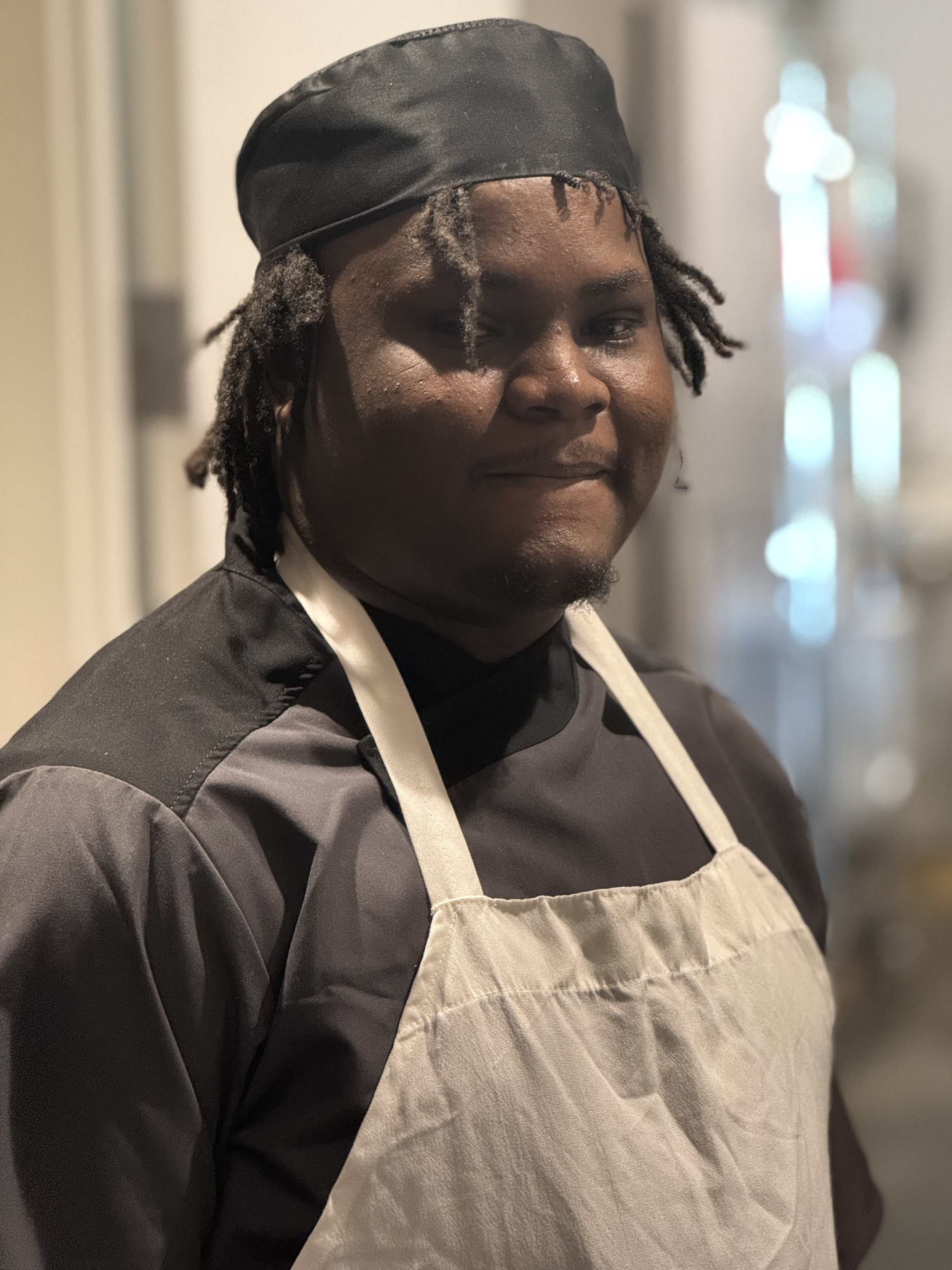 A person wearing a black chef's jacket, an off-white apron, and a black headscarf, standing indoors near blurred lights.