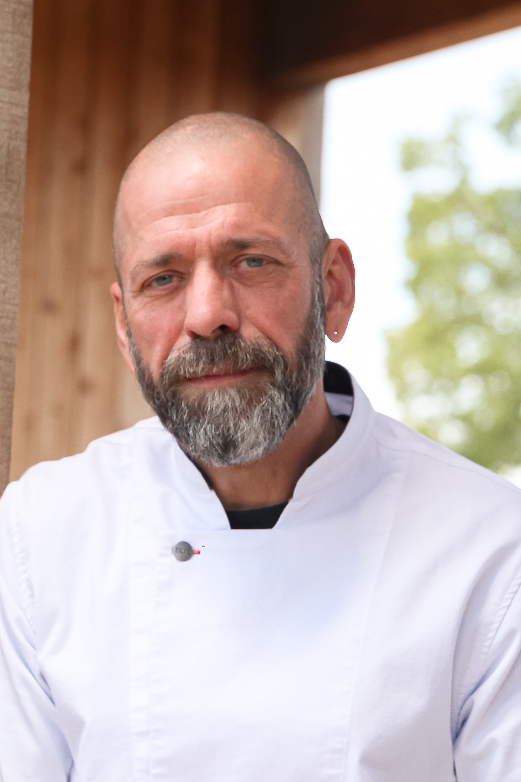 Close-up of a male chef with a shaved head, gray beard, and earring, wearing a white chef's coat, standing against a blurred background with greenery visible through an opening.