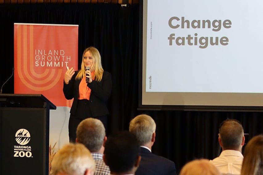 A woman giving a presentation at the Inland Growth Summit; a slide shows the words 'Change fatigue'; audience members are seated and listening.