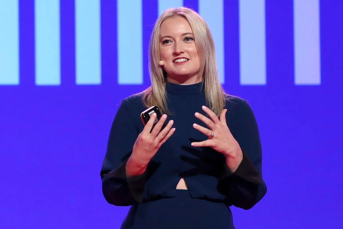 A woman with blonde hair, wearing a dark long-sleeve top, speaking on stage with a headset microphone and holding a smartphone in her right hand. She has her left hand on her chest and is smiling.