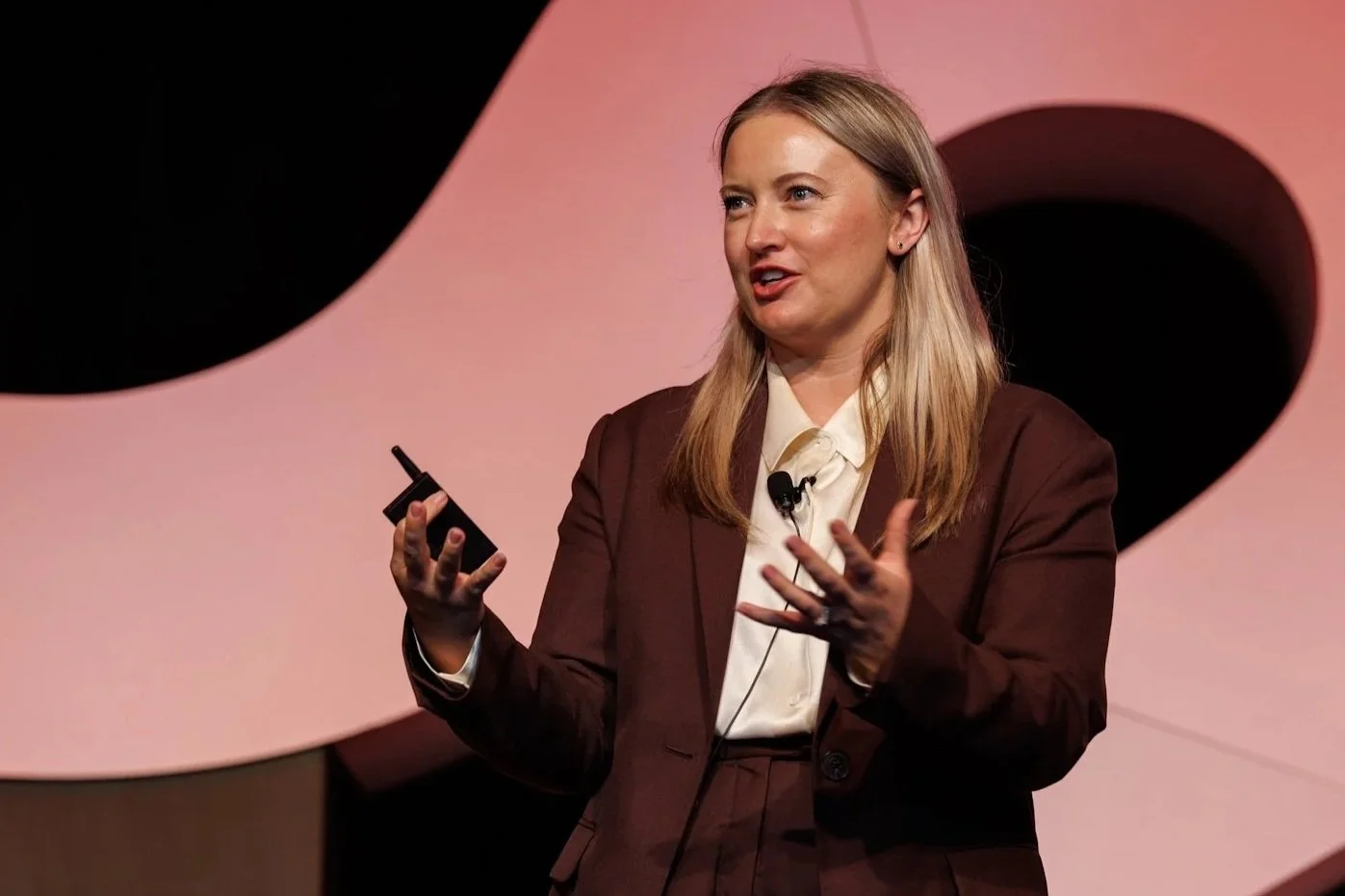 A woman with long blonde hair wearing a brown suit jacket and white blouse is speaking on stage, holding a remote control in her right hand and gesturing with her left hand. Behind her is a pink background with black and pink geometric shapes.