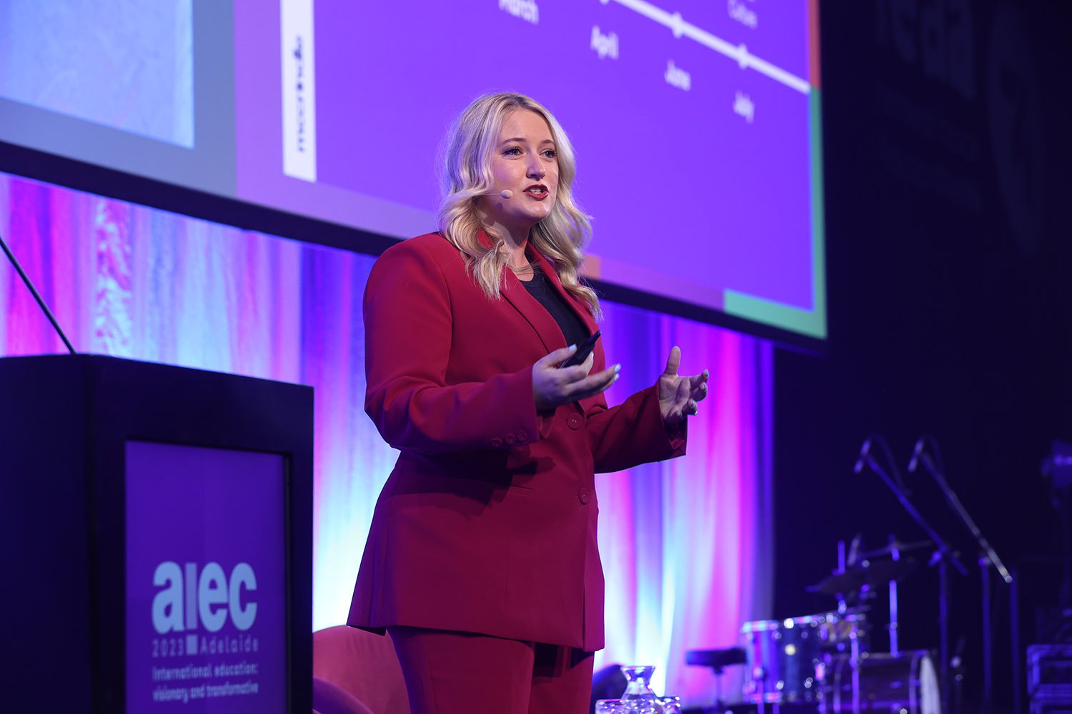 A woman with blonde hair wearing a red suit giving a presentation at a conference with a purple and blue background.