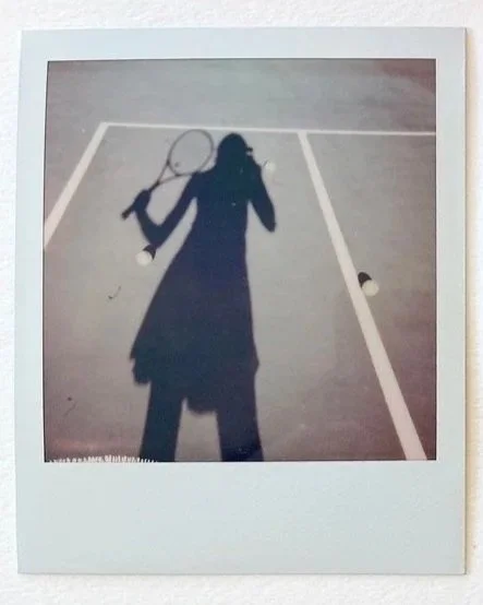 A polaroid of a woman's shadow as she holds a tennis racket on a tennis court.