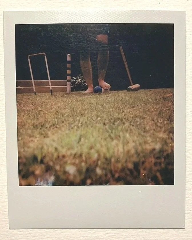 A polaroid of a person's legs and feet standing on a croquet lawn with a mallet and croquet balls.