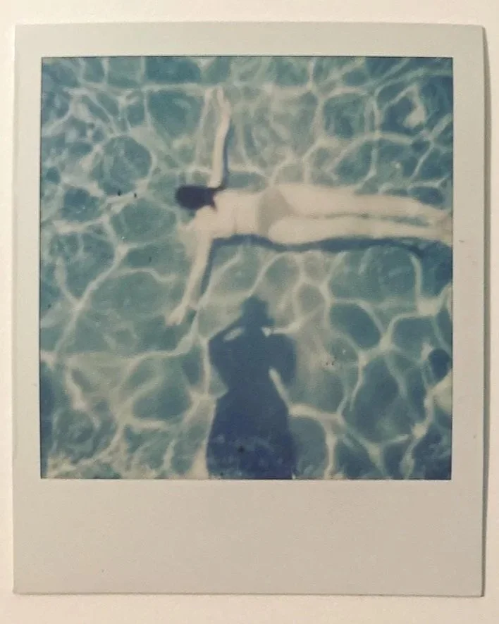 A polaroid of a woman swimming through a pool, with the photographer's shadow visible on the water.