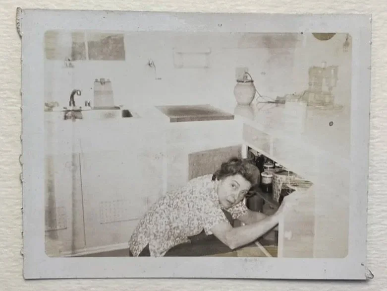 A photograph of a  woman crouching in a kitchen, reaching into an open cabinet.