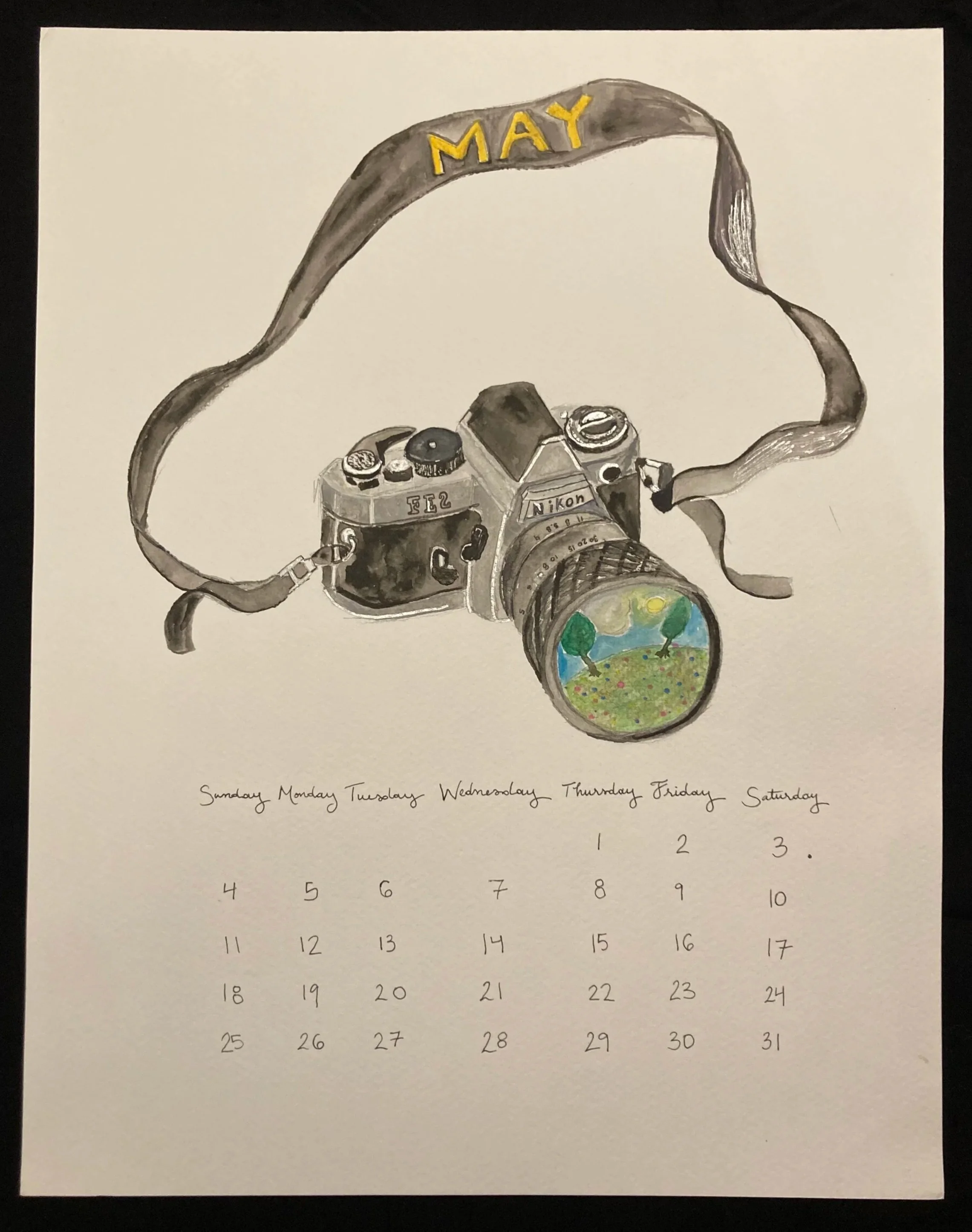 Calendar page for May, with a black and white illustration of a Nikon camera featuring a small painted scenic landscape with trees and a blue sky on the camera lens. The word "MAY" is written in yellow on a black camera strap.