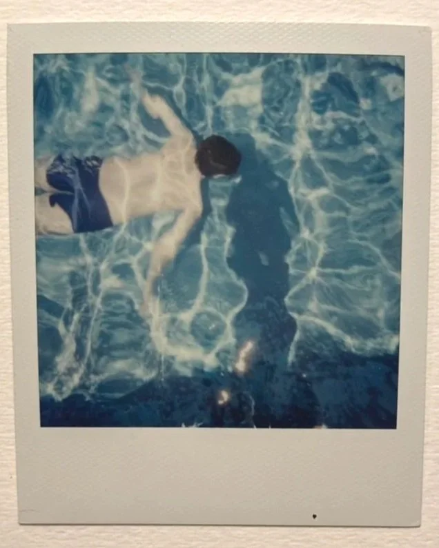 A polaroid of a man swimming through a pool, with the photographer's shadow on the water.