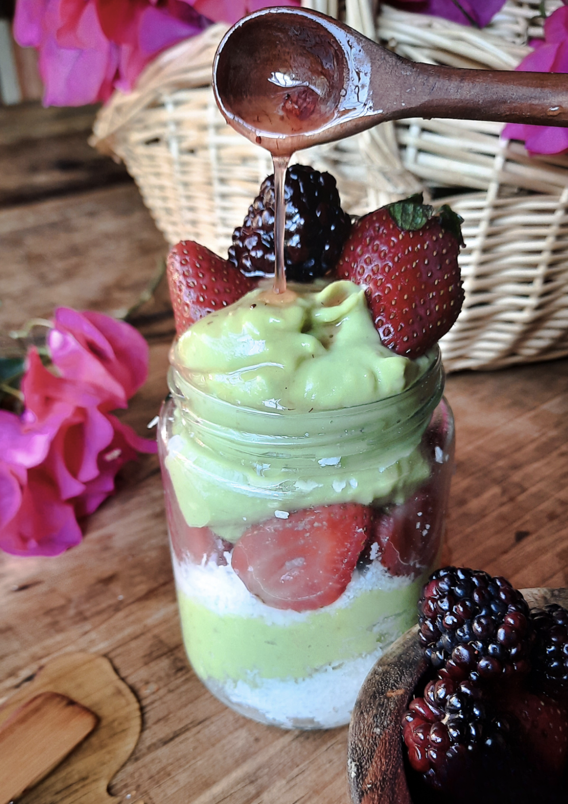 Verrine de dessert avec de la crème d'avocat, des fraises, des mûres et du parfum de miel, servie sur une table en bois avec des fleurs roses en arrière-plan.