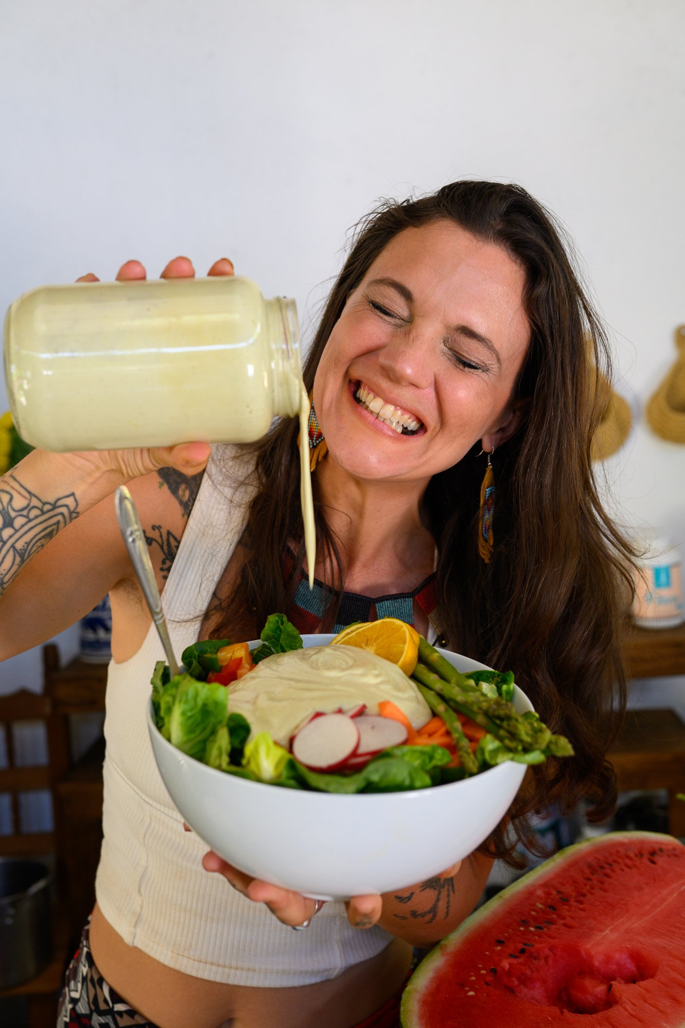 Une femme souriante verse de la vinaigrette sur une grande salade dans un bol, avec une pastèque coupée en premier plan.