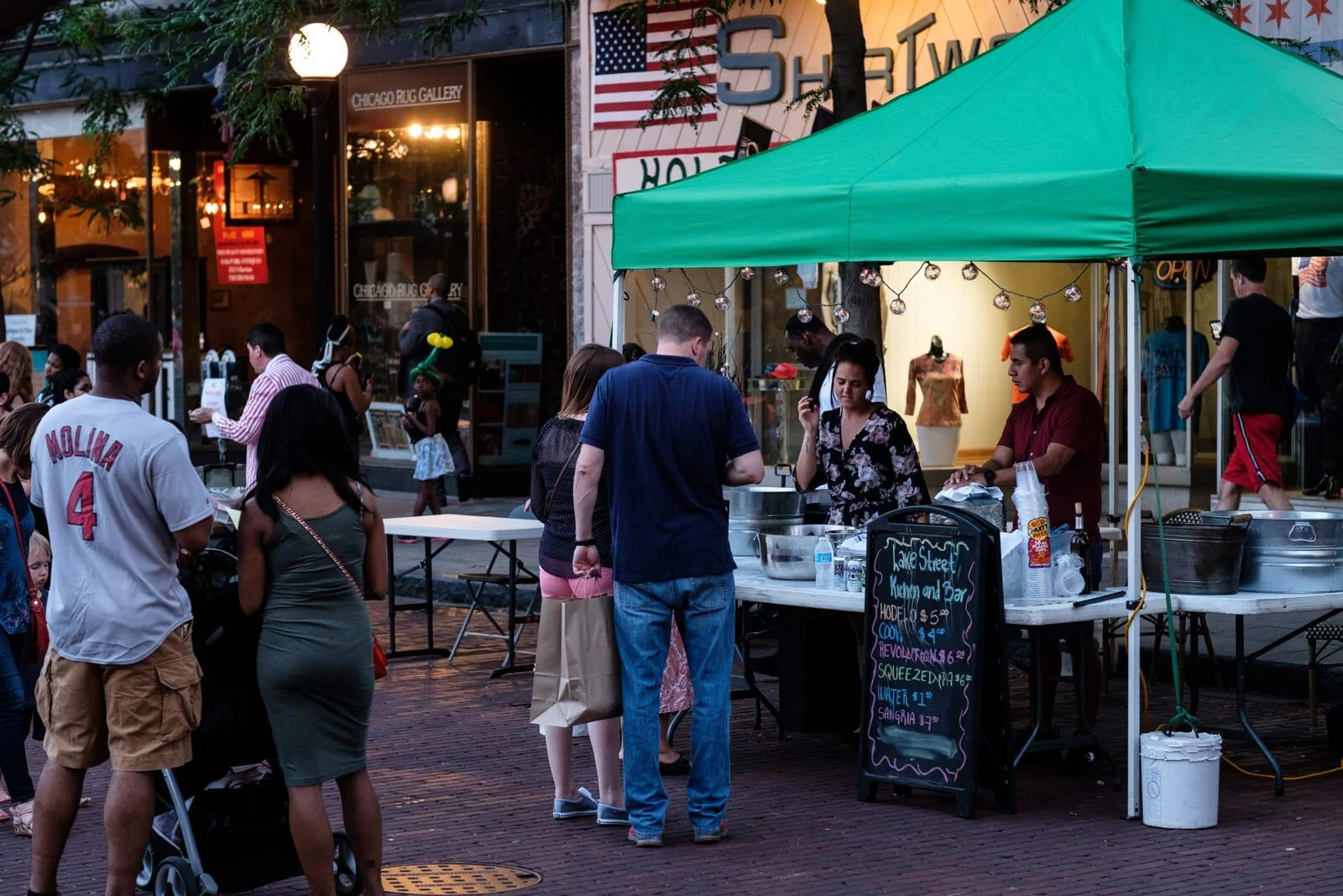 Street scene with a food stand under a green canopy and a crowd of people waiting and walking by. The stand has a chalkboard menu and is situated in front of a shop with mannequins and clothing inside. An American flag is visible on the building in the background.