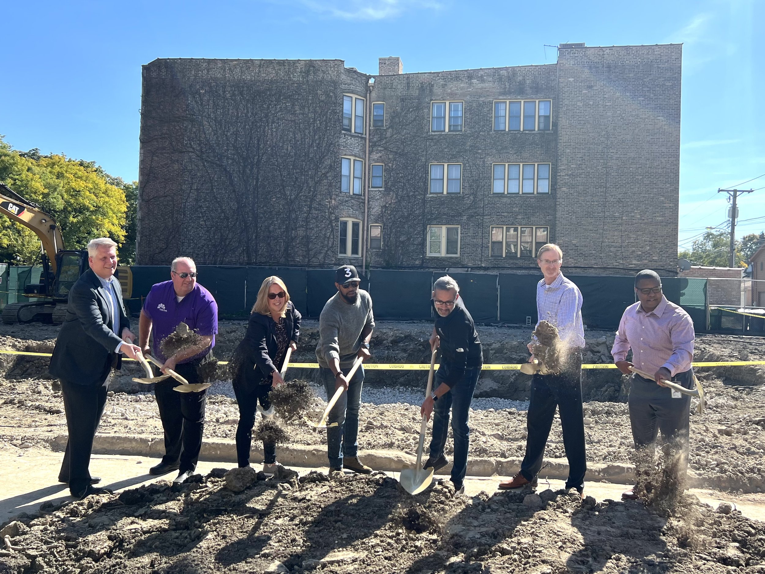 A group of people at a construction site with shovels, some smiling and working, with an urban building in the background under a clear blue sky.
