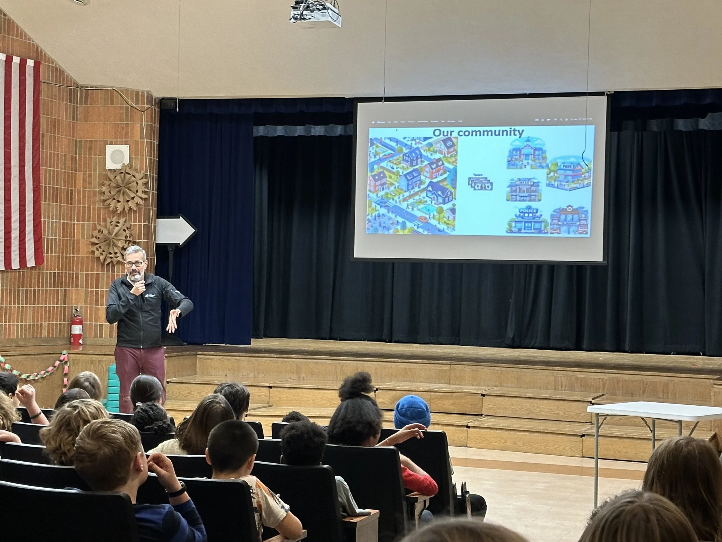 A man with glasses presenting to a large group of children seated in an auditorium. A presentation slide titled 'Our community' shows illustrations of houses, a park, a police station, and other community buildings.
