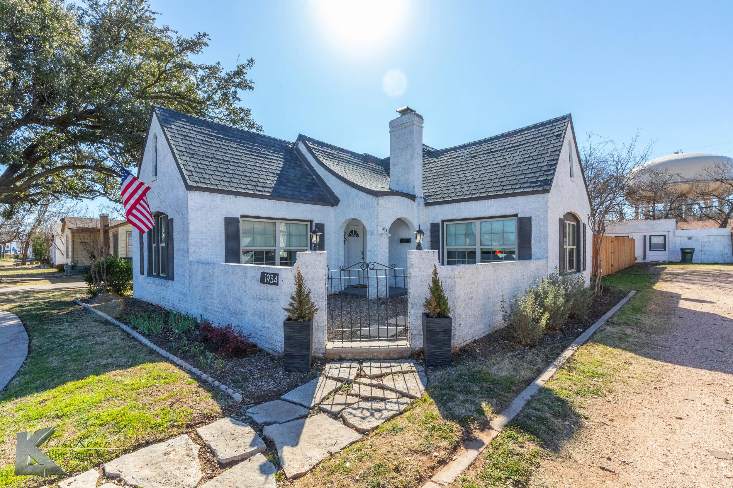 White brick house with black shutters, a front porch gate, and an American flag on the porch. The house has a steeply pitched roof with multiple gables. The house number is 1934. There's a large tree to the left and a water tower visible in the background to the right. The yard has a small garden with bushes and a sidewalk leading to the front door.
