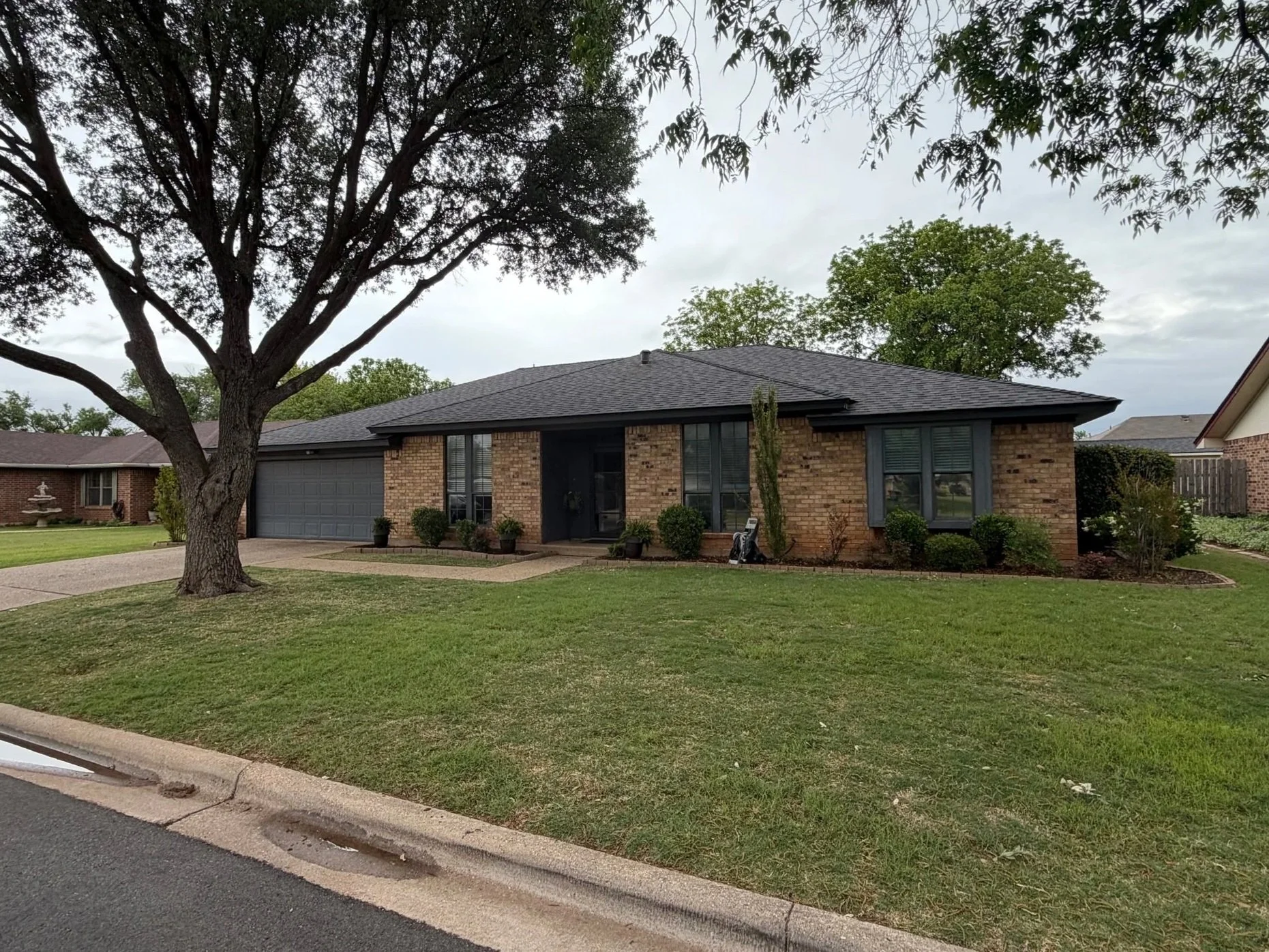 A brick house with a gray roof, black front door, and large windows, surrounded by green lawn, trees, and shrubs under a gray sky.