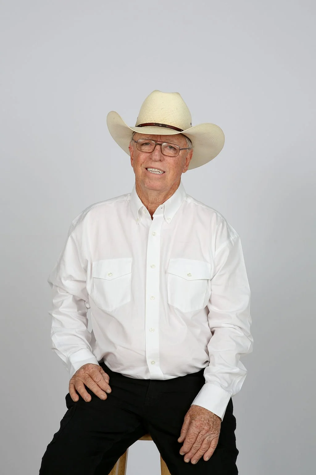 An elderly man with glasses wearing a cowboy hat, white button-up shirt, and black pants, sitting on a stool against a plain light gray background.