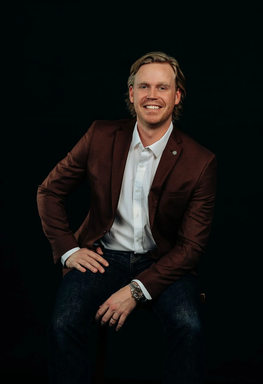 A smiling man with blond hair, wearing a white shirt, a maroon blazer, dark jeans, and a watch, posing against a black background.