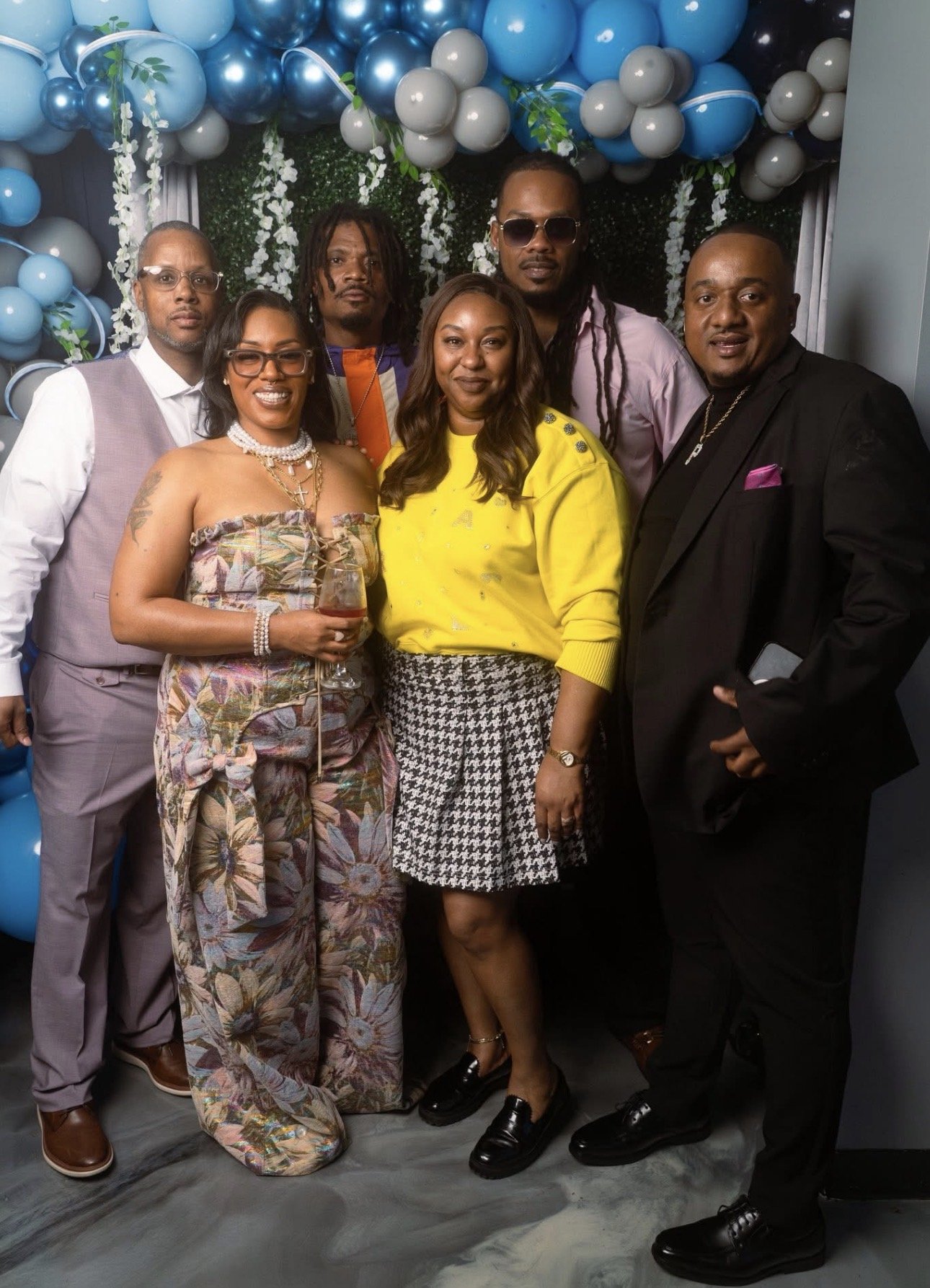 Group of seven people, five men and two women, posing together at a celebration with a backdrop of blue, white, and black balloons and green foliage. One woman is holding a drink, and some are wearing sunglasses and fashionable clothing.
