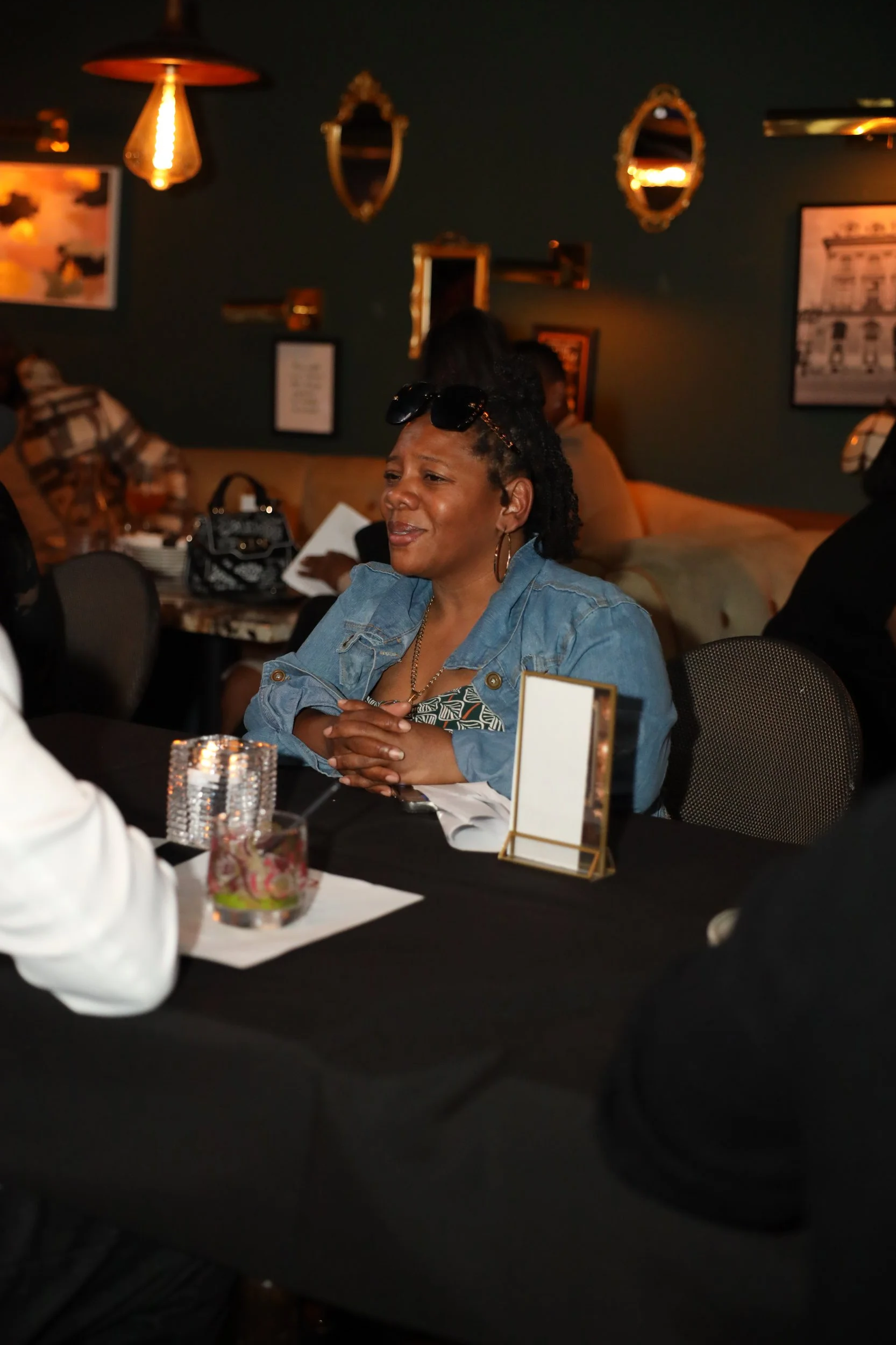 A woman with dark curly hair, wearing a denim jacket and sunglasses on head, sitting at a table in a dimly lit restaurant or bar, with a wall decorated with mirrors and framed pictures in the background.