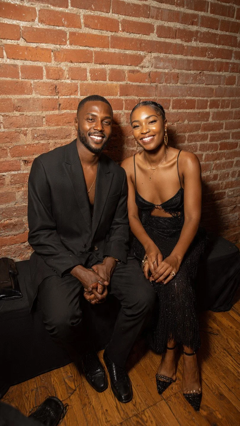 A smiling man and woman sitting closely together against a brick wall. The man is dressed in a black suit, and the woman is wearing a black dress with thin straps and sparkly details. They are sitting on a black bench or sofa, with a brick wall in the background.