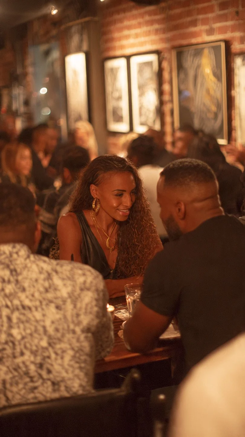 A woman with curly hair wearing gold jewelry smiling and talking to a man with a beard in a dimly lit, lively restaurant decorated with framed artwork and exposed brick walls.