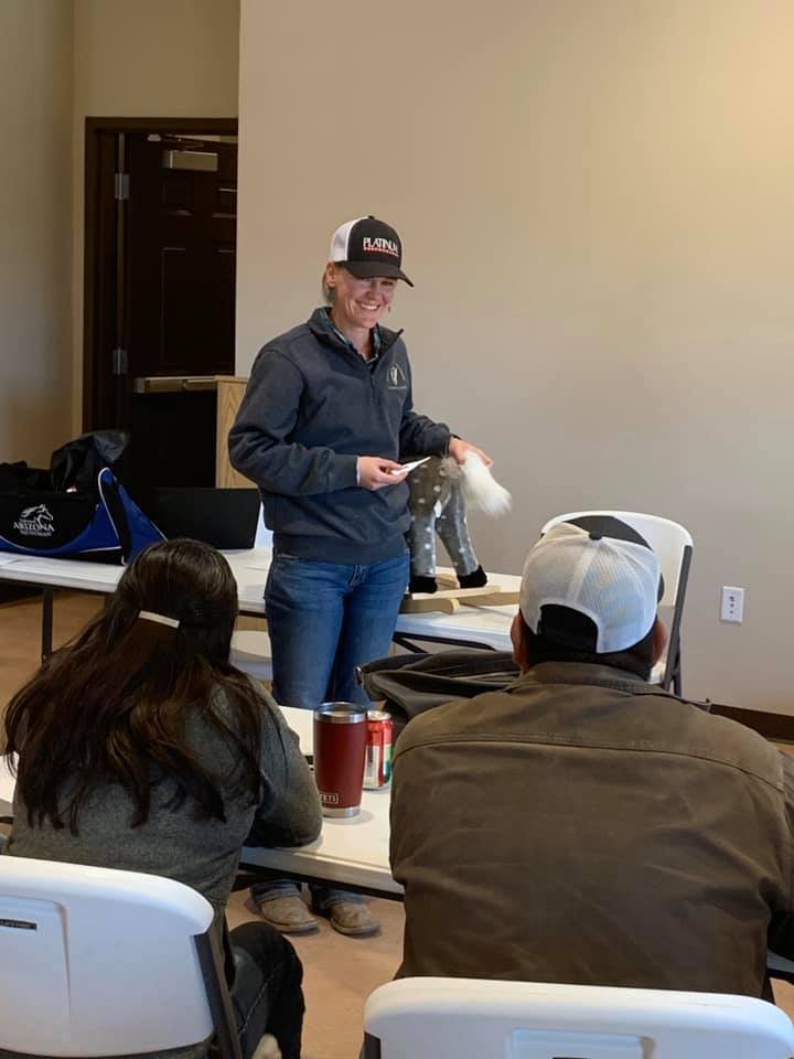 A woman standing and smiling, wearing a dark jacket, jeans, and a trucker hat, holding a paper and a dog toy, in a room with people seated at tables, including a man with a white cap and a woman with long dark hair.