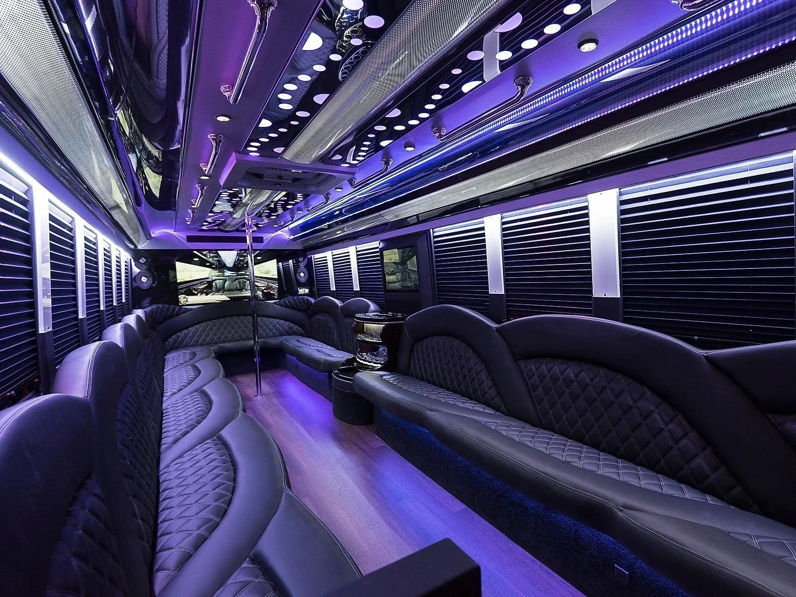 Interior of a luxurious party bus with purple ambient lighting, leather seating, and a dance pole. The design includes patterned seats and a sleek ceiling with LED lights.