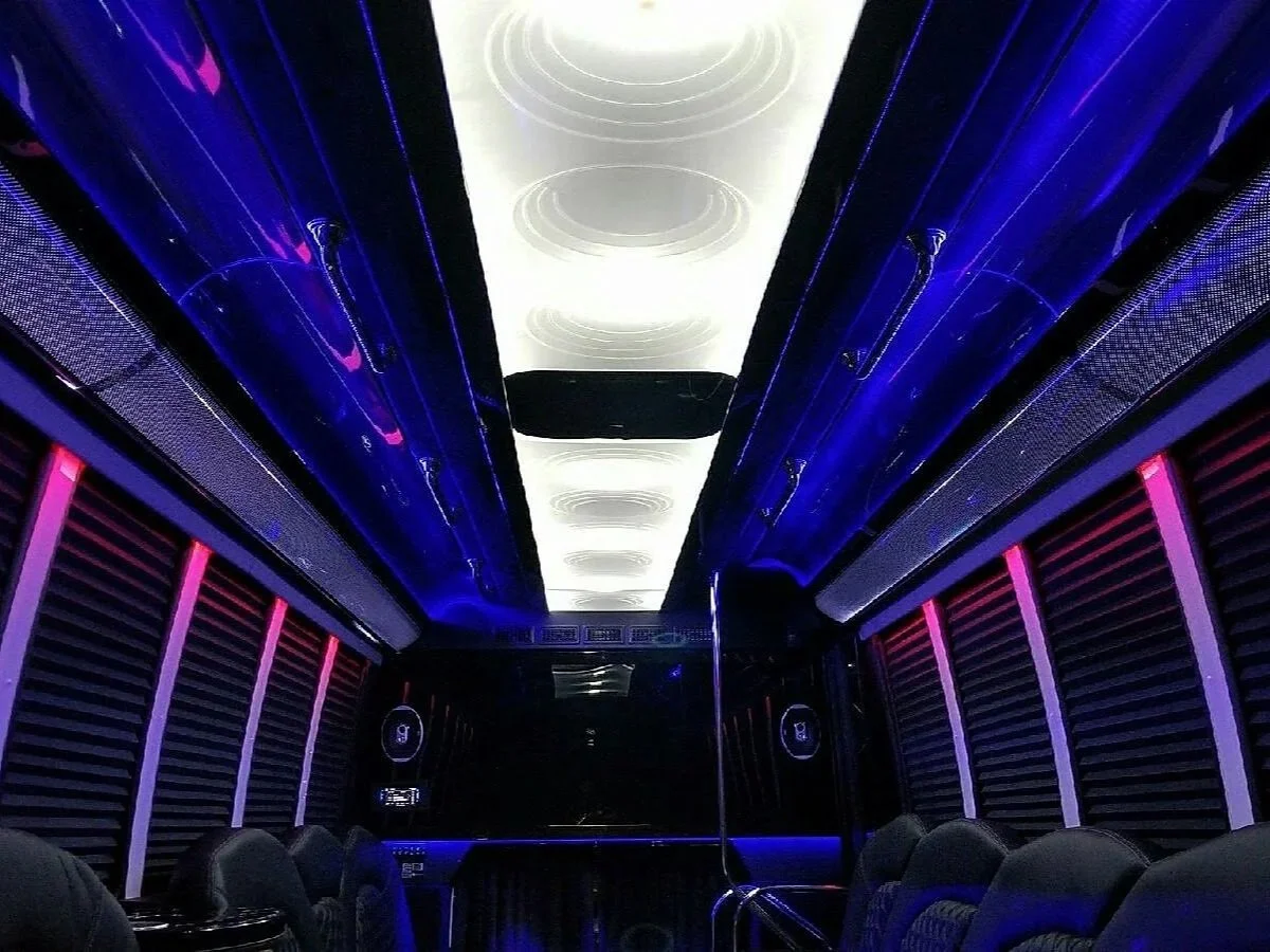 Interior of a party bus with blue and pink LED lighting, lounge seating, and ceiling speakers.