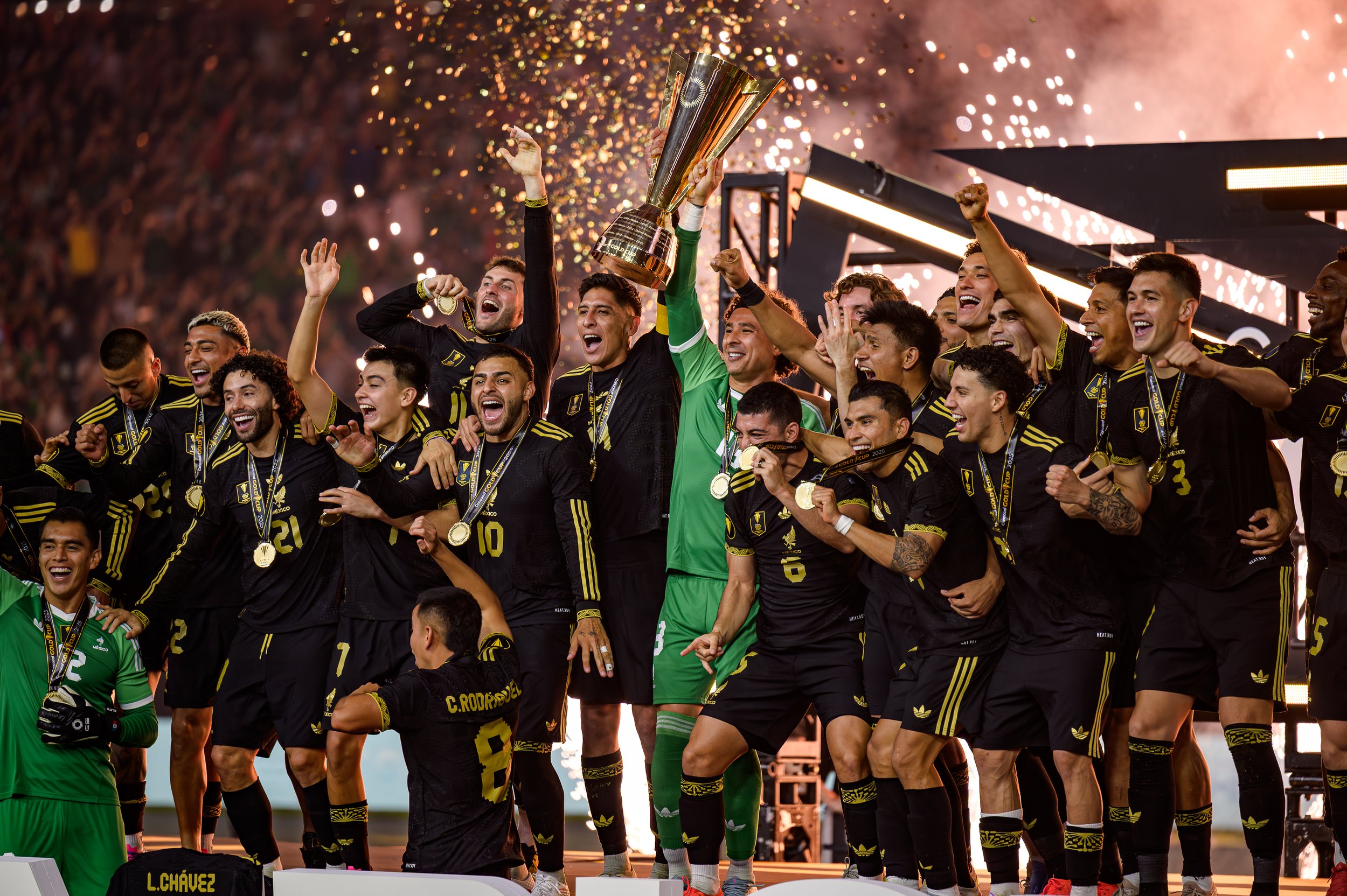 A team of soccer players celebrating their victory, holding a large trophy, wearing medals, and cheering amidst fireworks and a crowd.