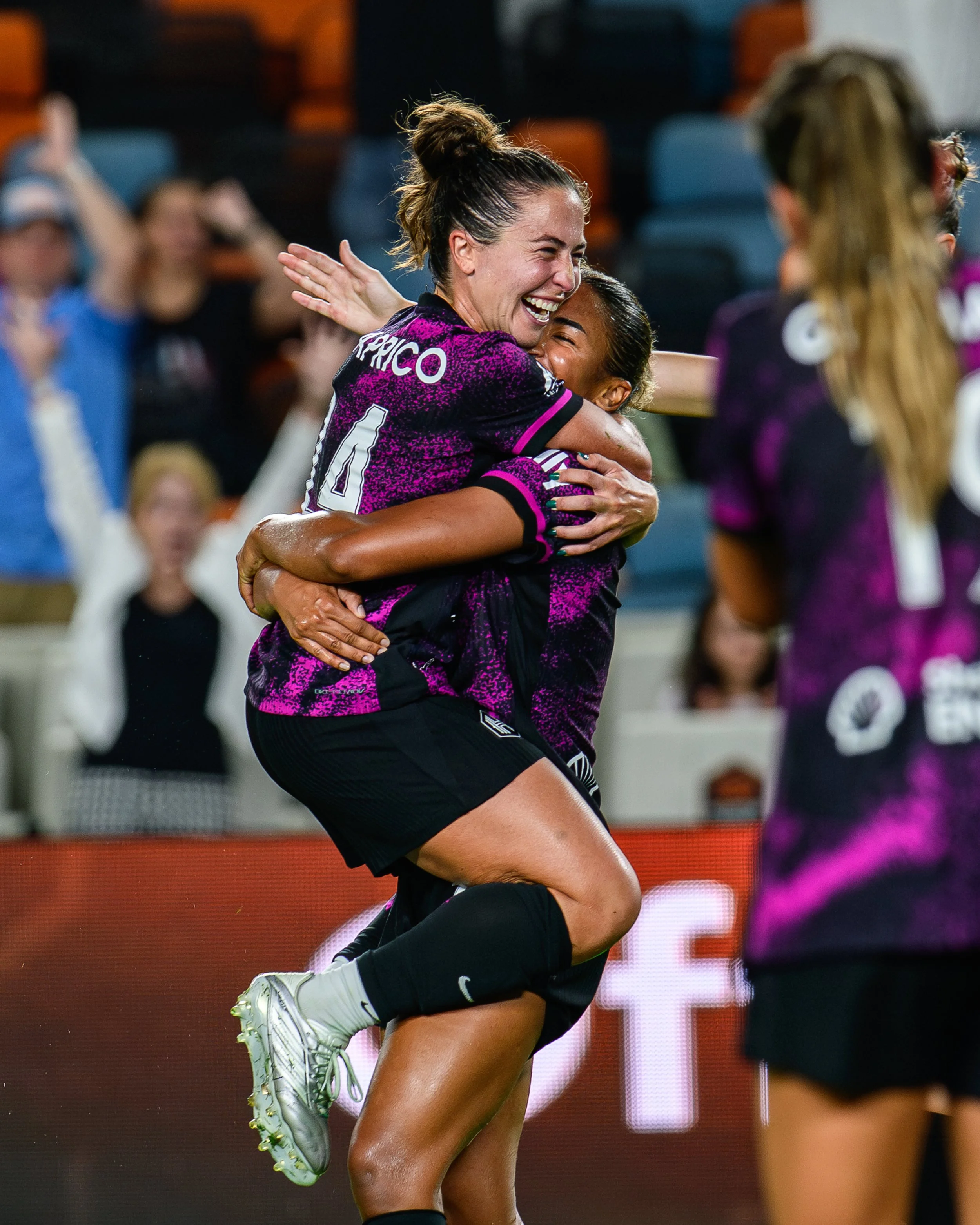 Two female soccer players in purple jerseys celebrating a goal, one player is lifting and hugging the other with a joyful expression on her face, surrounded by blurred spectators.