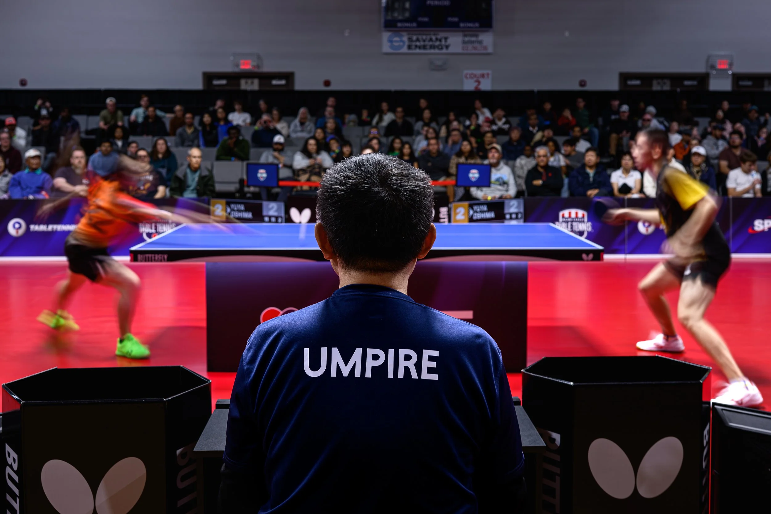 A table tennis match with two players in action, one in an orange shirt and black shorts, the other in a black and yellow shirt and black shorts, on a red table tennis court. An umpire with a navy blue shirt marked 'UMPIRE' sits in front, with a larg