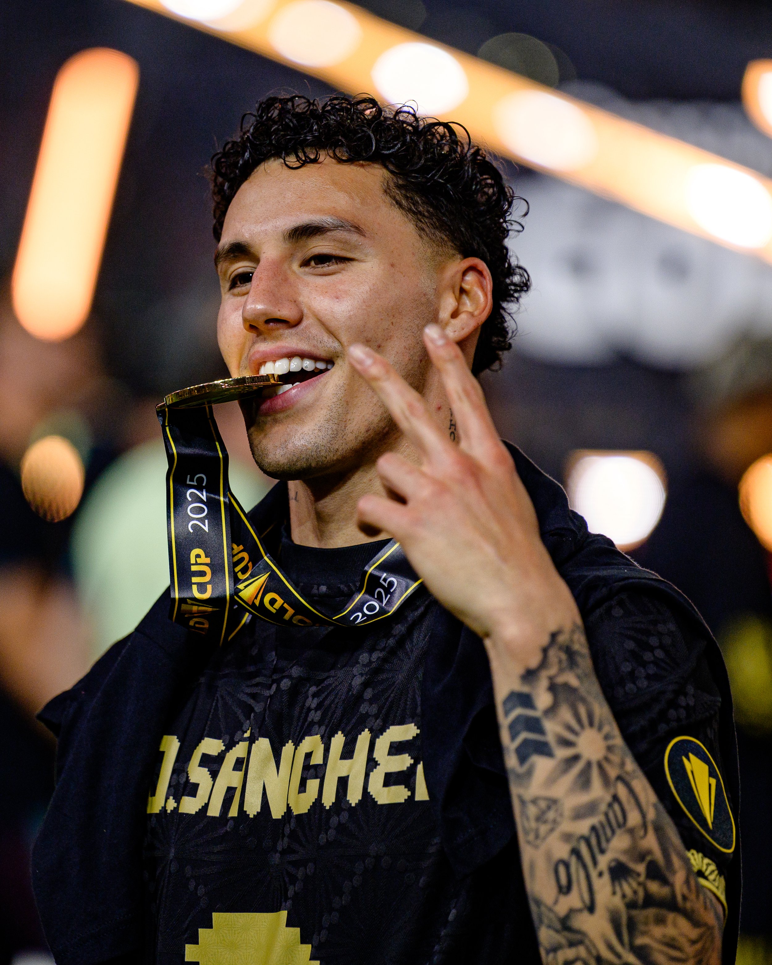 A young man with curly dark hair and tattoos on his arm smiling as he bites a gold medal hanging around his neck, with a blurred background of bright lights.