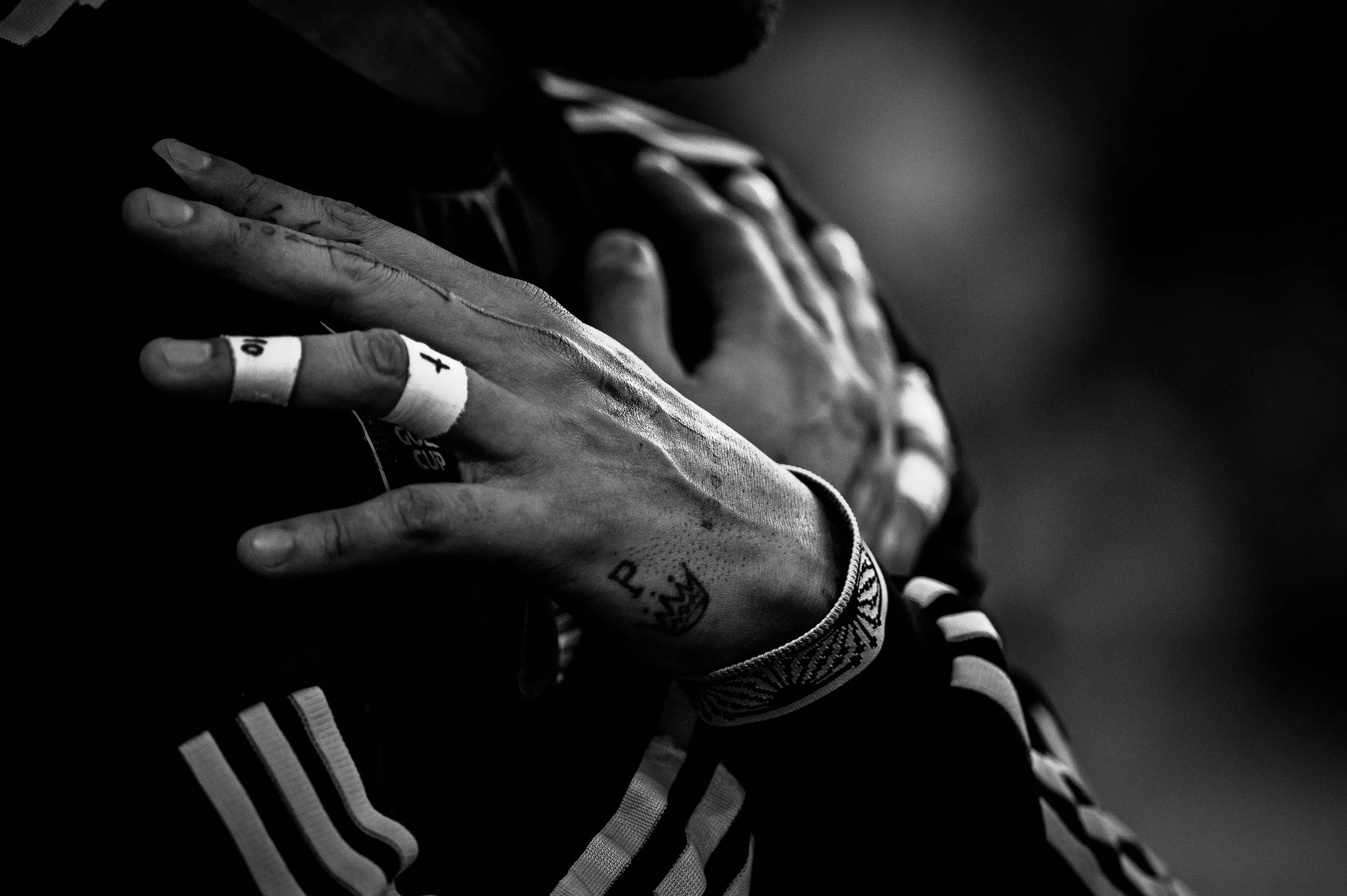 Close-up black-and-white photo of a person's hands, with one hand resting on their chest. The hands have tattoos and are wearing wristbands. The person is dressed in a sports jacket with stripes.
