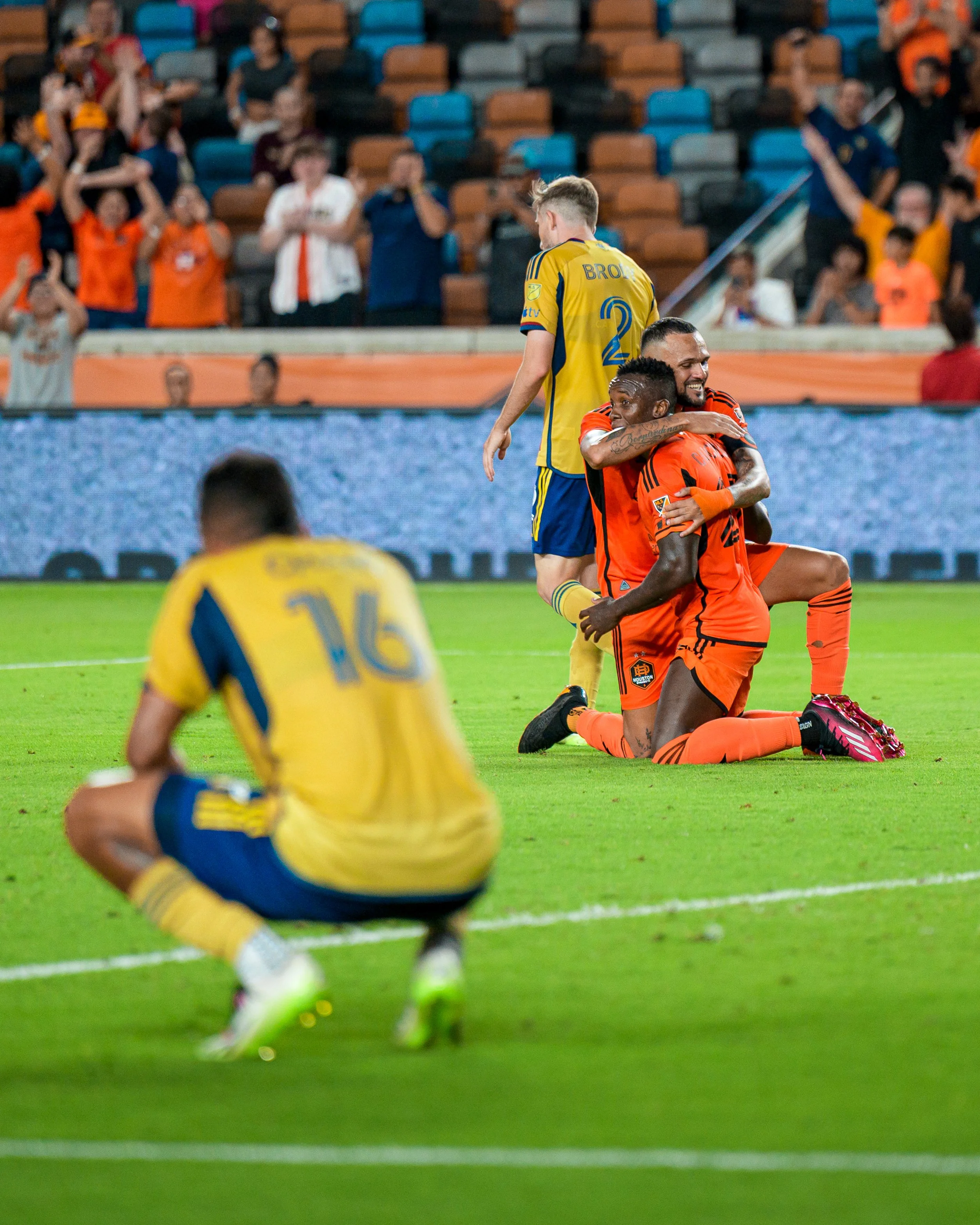 Soccer players celebrating on the field, with one kneeling and hugging another, while a third player sits on the ground. Fans are cheering in the background.