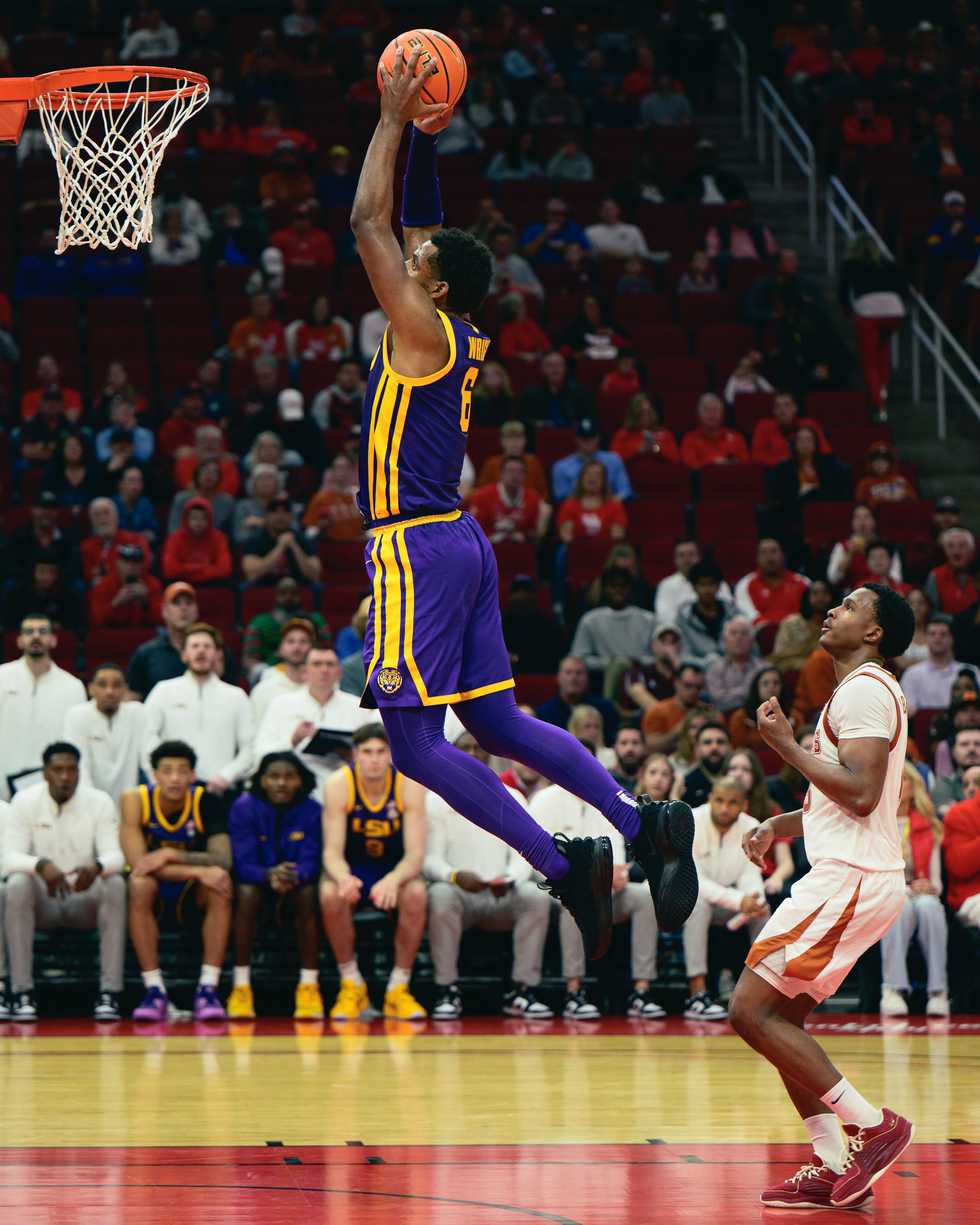 A basketball player in a purple and yellow uniform is jumping in the air to shoot the ball towards the hoop, with an opposing player in a white and orange uniform watching nearby, during a game in an indoor stadium with a crowd of spectators in the b