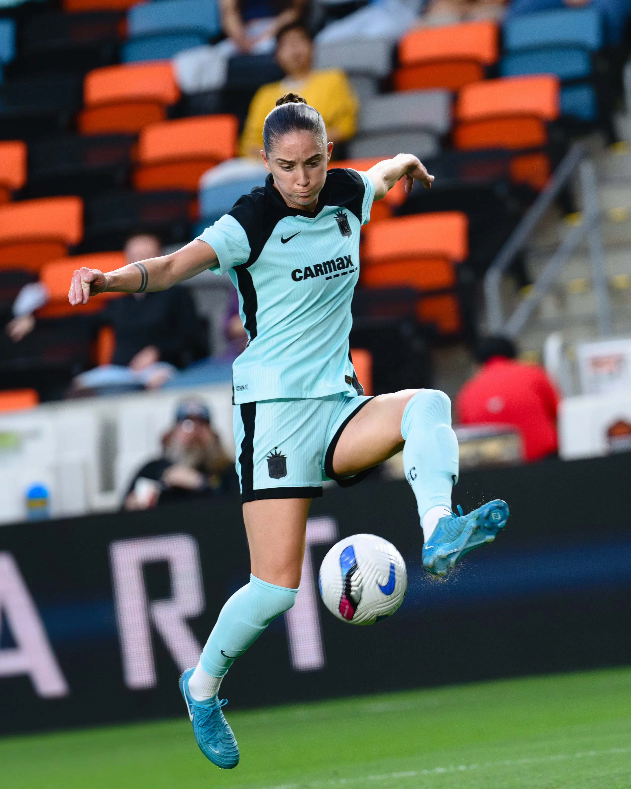 A female soccer player in a light blue uniform with black accents, mid-air kicking a soccer ball during a game.
