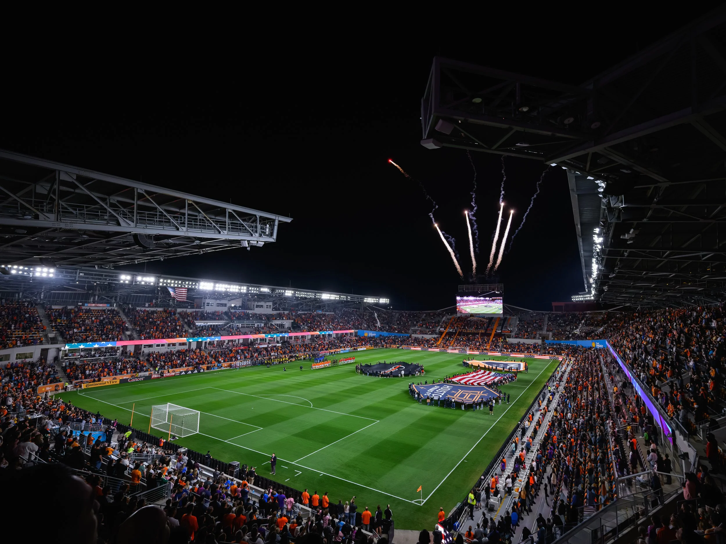 Nighttime football stadium filled with spectators, fireworks in the sky, and flags on the field, during a celebratory event.