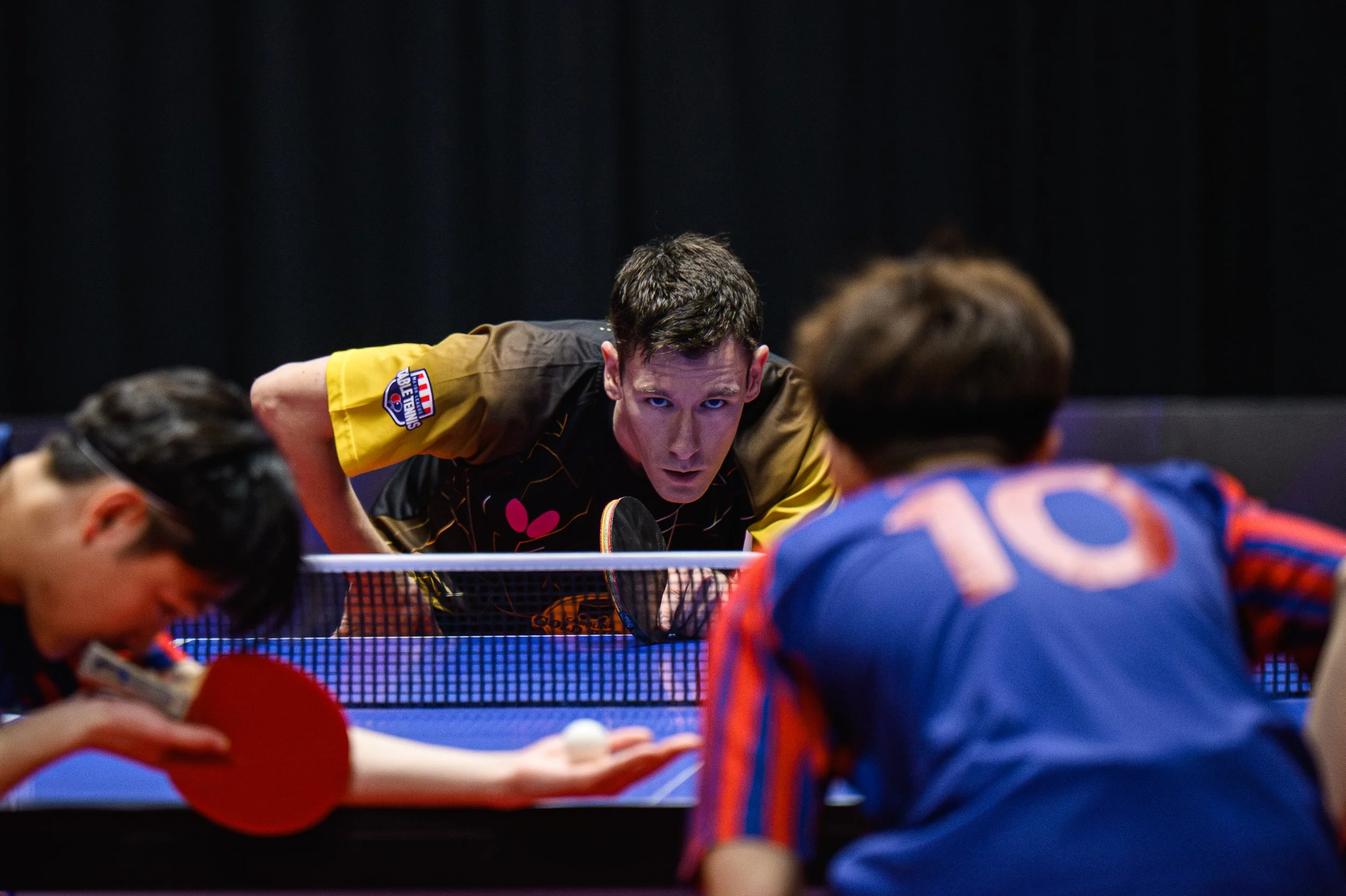 Two boys playing table tennis with a woman, with one boy wearing a black and yellow shirt and the other boy wearing a blue and orange shirt, and a man in the background.