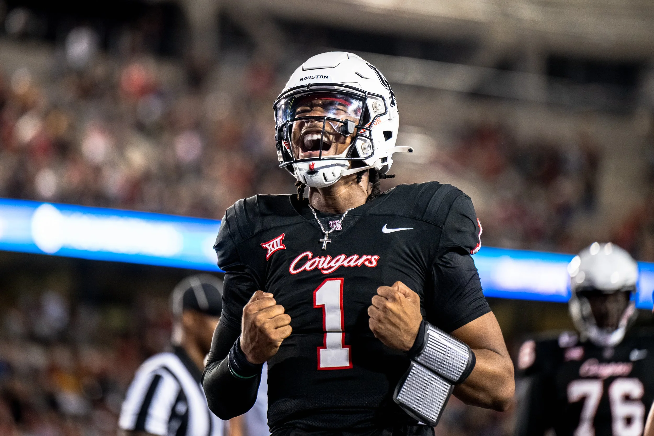 A college football player wearing a black jersey with 'Cougars' and the number 1, celebrating with clenched fists and an open mouth, wearing a white helmet and wristbands, on the field with a blurred crowd in the background.