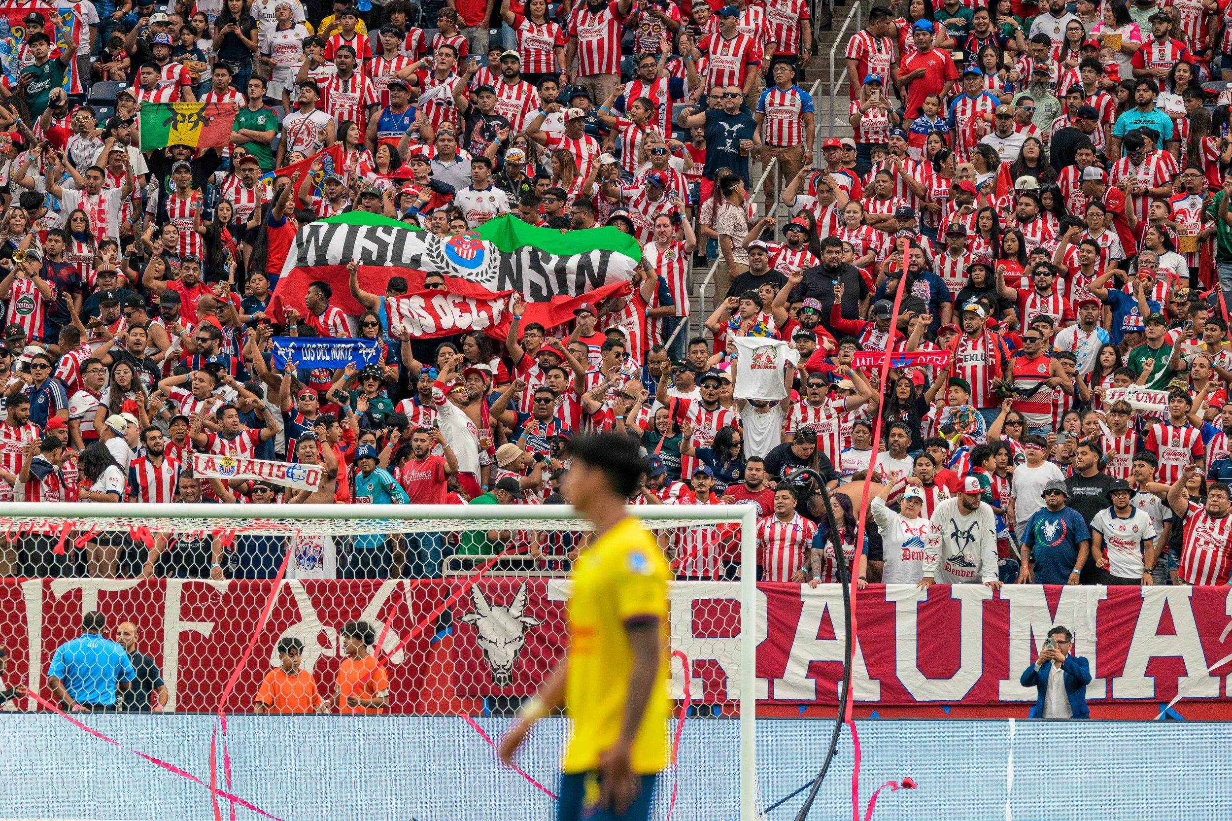 Crowd of soccer fans in red and white striped jerseys at a stadium, cheering and holding banners. A blurred player in a yellow jersey is in the foreground near the goal, with a photographer taking pictures.