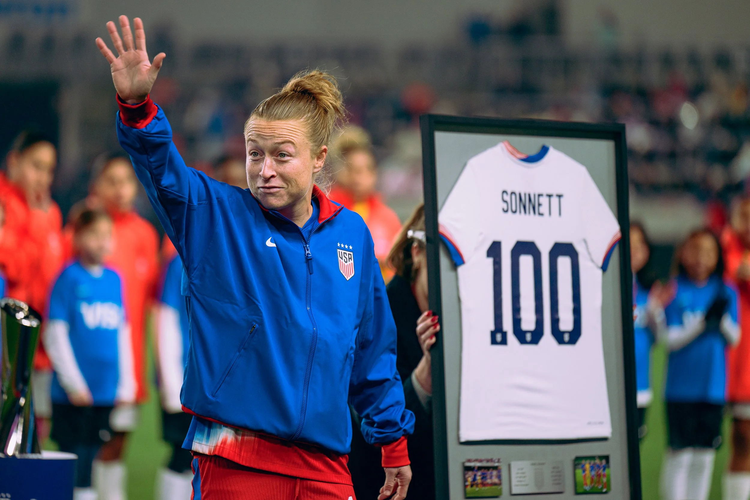 A woman in a blue USA soccer jacket waving, standing next to a framed soccer jersey with the name Sonnett and the number 100, at a sports event with other players and spectators in the background.