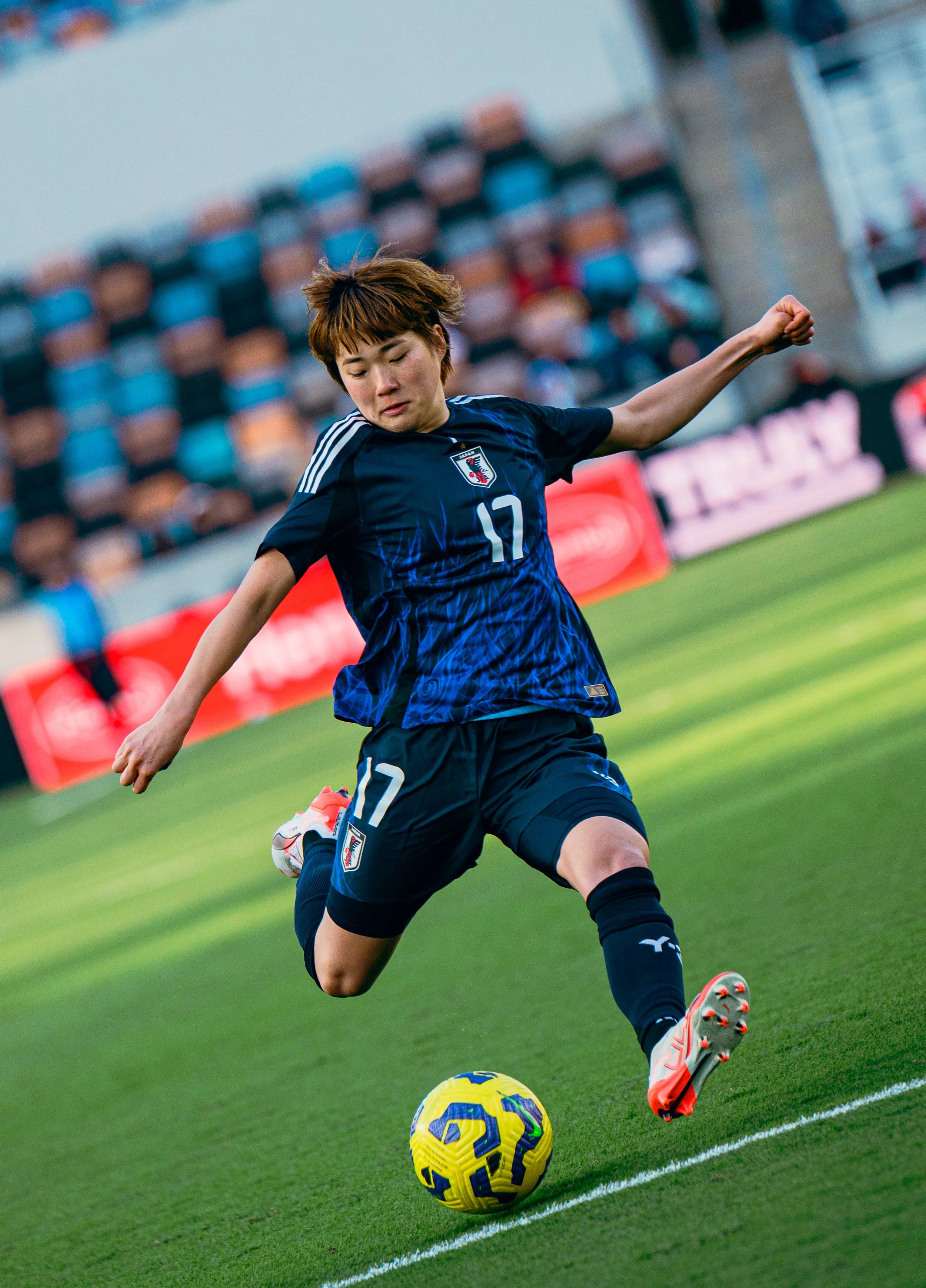 A female soccer player in a dark blue jersey with the number 17, kicking a yellow and purple soccer ball on the field.