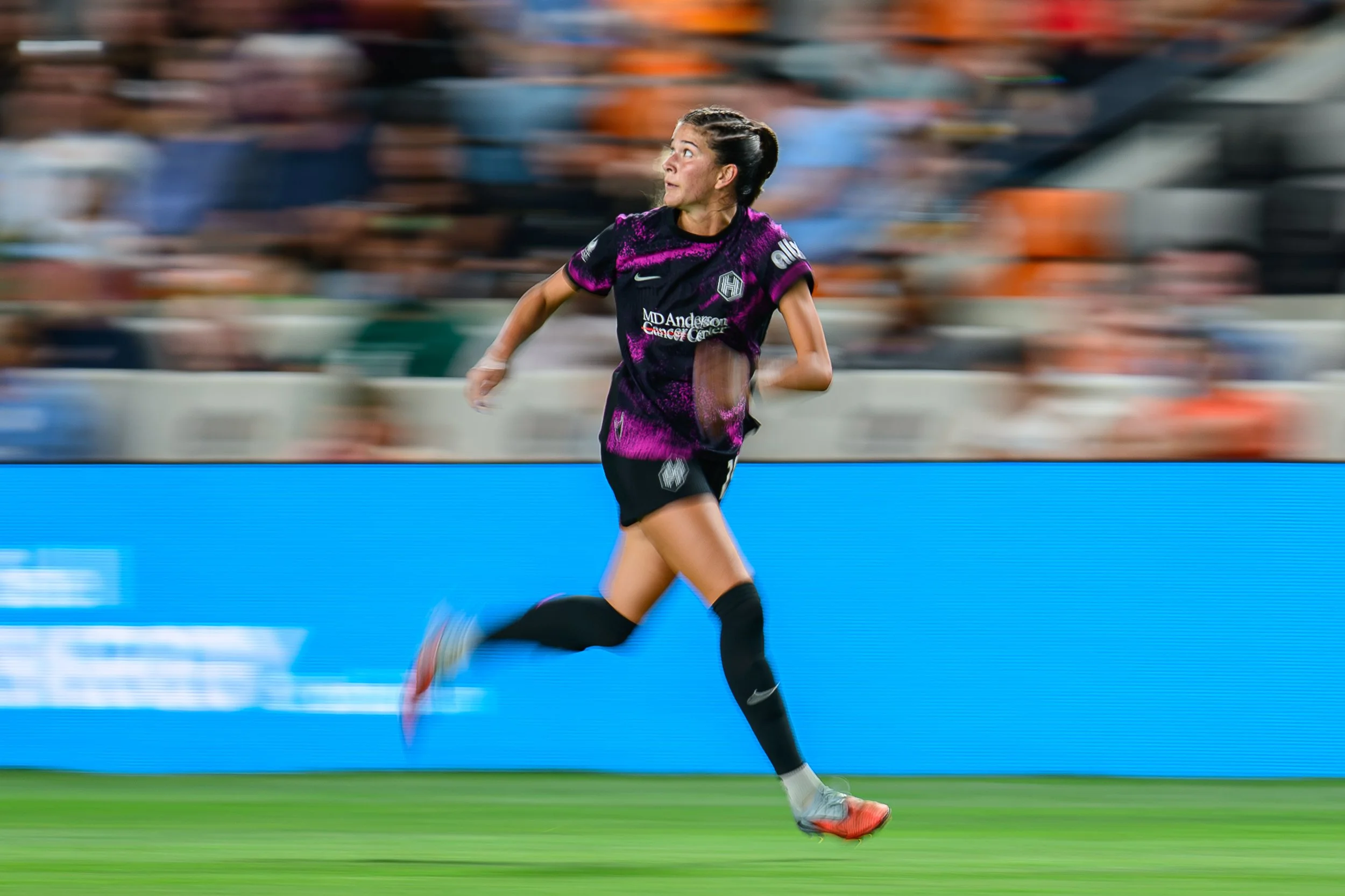 A female soccer player running on the field during a match, wearing a black and purple uniform with logos, with a blurred crowd in the background.