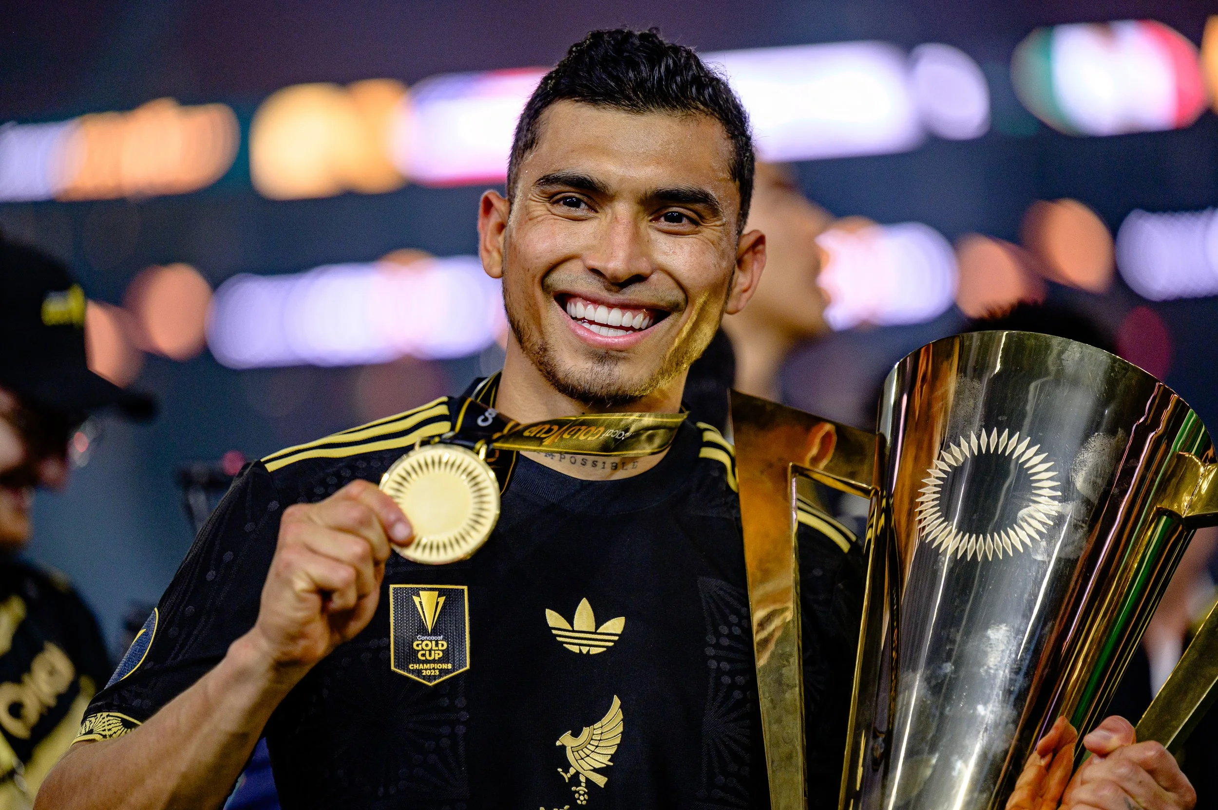 A man in a black and gold sports jersey holding a gold medal and a large trophy, smiling after a victory.